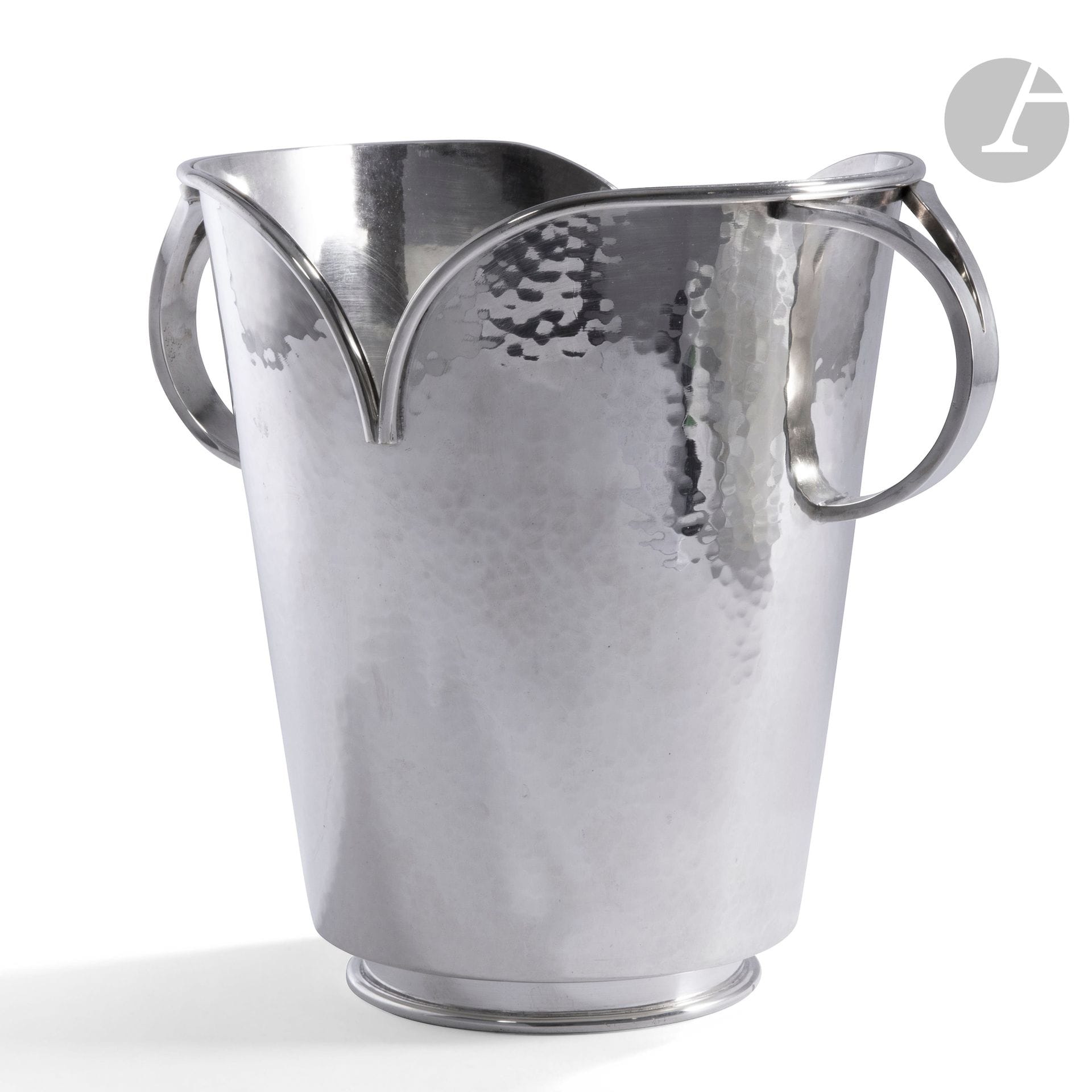 JEAN DESPRÉS (1889-1980)
Découpe cordiforme
Très rare et élégant seau à Champagne (rafraîchissoir) en métal argenté à surface martelée. Le corps évasé repose sur un talon lisse en retraitreçu par une base circulaire; la bordure en découpe titre et rehaussée d’un jonc cylindrique en ressaut. Les anses curvilignes et formant chacune un Y; leur surface traitée lisse. Réalisation des années 1970. Porte le poinçon de Maître Orfèvre sur le pourtour de la base. H.24,5cm – L.28cm