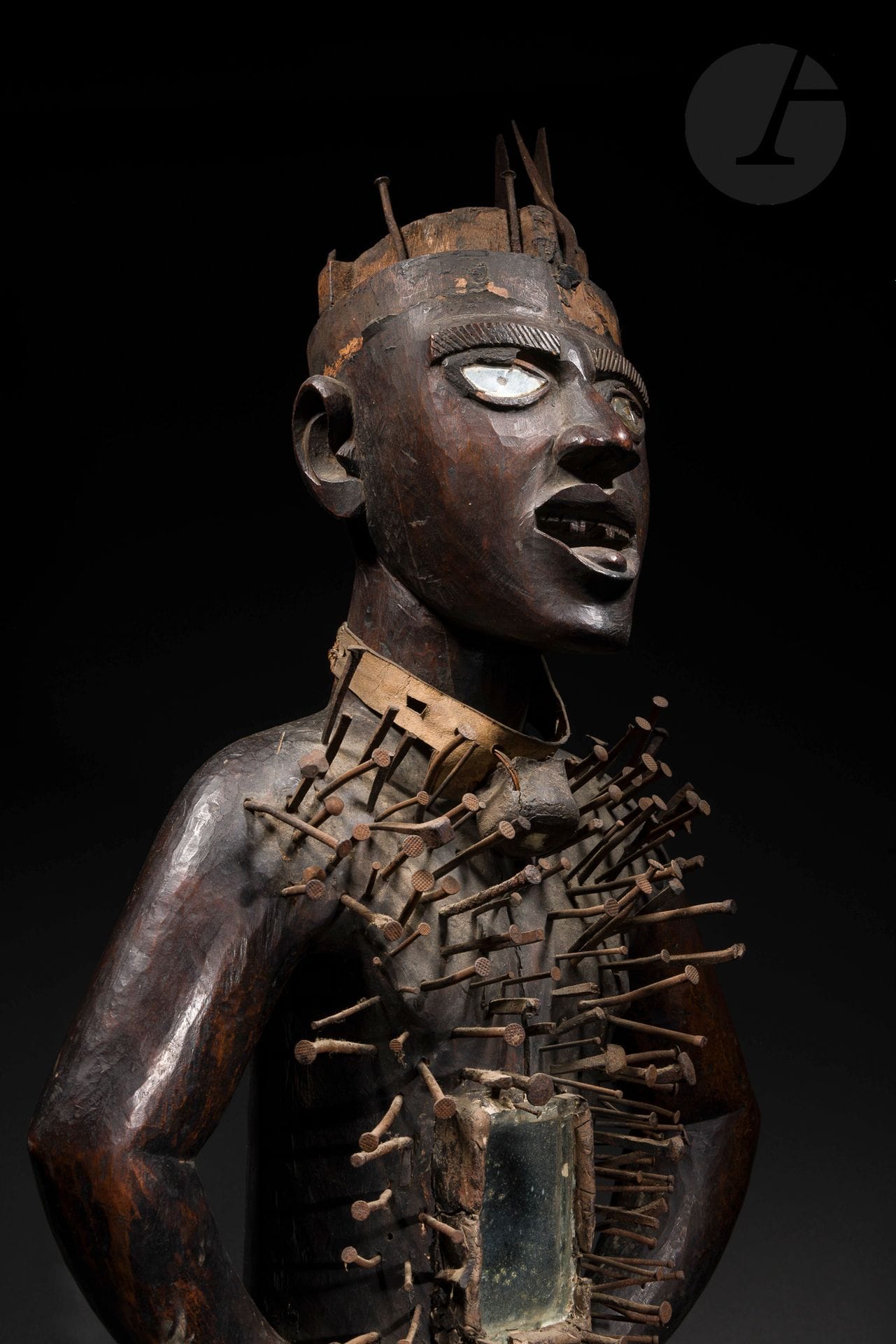 Un important et ancien «fétiche à clous» Nkisi Nkonde (ou Nkisi Nkondi). 
Les grandes statues appelées par les occidentaux «fétiches à clous» font de toute évidence partie des objets parmi les plus emblématiques, mythiques (voir mythologique), et terrifiant ou pour le moins inquiétant de tous les arts d’Afrique. La redécouverte d’un grand Nkisi Nkonde inédit constitue forcément une certaine émotion, un petit événement, qui mérite qu’on évoque au moins un peu leurs origines et leurs fonctions.
Dans l’ancienne tradition Bakongo, dont l’ancien royaume fût découvert en 1482 par le navigateur portugais Diego Cam, et qui s’effrita jusqu’à disparaître à la fin du XIXe siècle, les statues Nkisi Nkondi sont des objets réputés puissant qui créent un pont entre le réel et l’irréel, le matériel et l’immatériel. 
Ils ont de multiples pouvoirs, des fonctions dans le cadre de pratiques thérapeutiques ou prophylactiques, capables d’actes annulant les effets néfastes de la sorcellerie, tenant à distance les maléfices, mais aussi comme «les chasseurs aguerris» ils peuvent «tendre des embuches aux menteurs, aux voleurs, aux adultères, à toutes sortes de personnes pouvant nuire aux structures de la société». En effet le mot Nkondi vient du verbe nkonda qui veut dire chasser, mais selon les traductions et la prononciation variant d’une région à l’autre cela peut vouloir aussi dire: alliance/accord/établir un serment. 
Le Nganga est le gardien qui veille sur le Nkisi Nkondi. Il a participé pleinement à sa création, et il est de fait en partie dépositaire de sa force et de ses pouvoirs. C’est entre le Nganga, qui fait donc office de guérisseur, devin, mais aussi homme de pouvoir judiciaire et politique, et ses «patients» ou ses «clients», que ces liens s’établissent, et les vœux, les prières, ou les demandes dont les clous, qui sont souvent frottés en terre et léchés avant d’être planter par le nganga lui-même, sont des témoins qui scellent cette alliance ou ce serment.
La position debout, les deux mains bien posées sur les hanches et les coudes largement dégagées du corps, avec la tête droite légèrement inclinée vers l’avant, se dit pakala qui signifie dresser l’oreille et symboliserait l’attente de mambu dans des affaires sérieuse ou judiciaire, avant d’attaquer de front. 
Une grande majorité des Nkisi Nkonde sont sculptés asexués même si certains sont clairement féminins ou masculins (environ 5%) ou même hermaphrodite (0,8%), la grande majorité (75%) des statues Nkisi Nkondi sont clairement sculptées sans indication de leur sexe, et ne représente donc à priori pas un personnage précis, ou un ancêtre. Un sanctuaire dédié leur était réservé, mais il semble aussi qu’ils aient pu servir de gardien sur les tombeaux de personnes importantes.
La présence ici de nombreux et anciens clous en fer forgés (en plus des clous d’importations d’époques coloniales), son style et son iconographie (bouche ouverte, dents taillées, langue tirée, belles oreilles bien sculptées), témoignent d’un art Bakongo d’une tradition encore bien ancienne, comme en attestent d’ailleurs aussi sa très belle patine et les reliquats de résine des anciennes charges reliquaires magiques bilongo qui ont depuis disparues.
Le Nkisi Nkondi de la collection Alice et Henri Dupont vient évidemment enrichir l’inventaire d’un corpus, somme toute, assez restreint de statues parmi les plus mythiques des arts d’Afrique, et s’impose au-delà de sa taille par une très belle et réelle puissante présence. 
Voir: Lehuard, Fétiches à clous du Bas- Zaïre, Arnouville 1980 et Musée Dapper, «Magies», Paris 1986.
Kongo (probablement Yombe), République du Congo ou Démocratique du Congo, XIXe siècle
Bois, métal, clous et fer forgé, verres, résine et reliquats, cuir, miroirs (dont un refixé sans la charge), accidents visibles, fentes et érosions d’ancienneté, très belle et ancienne patine d’usage.
H.: 88cm

Provenance:
Collection Alice et Henri Dupont, Lille, acquis c. 1960
Transmis par descendance