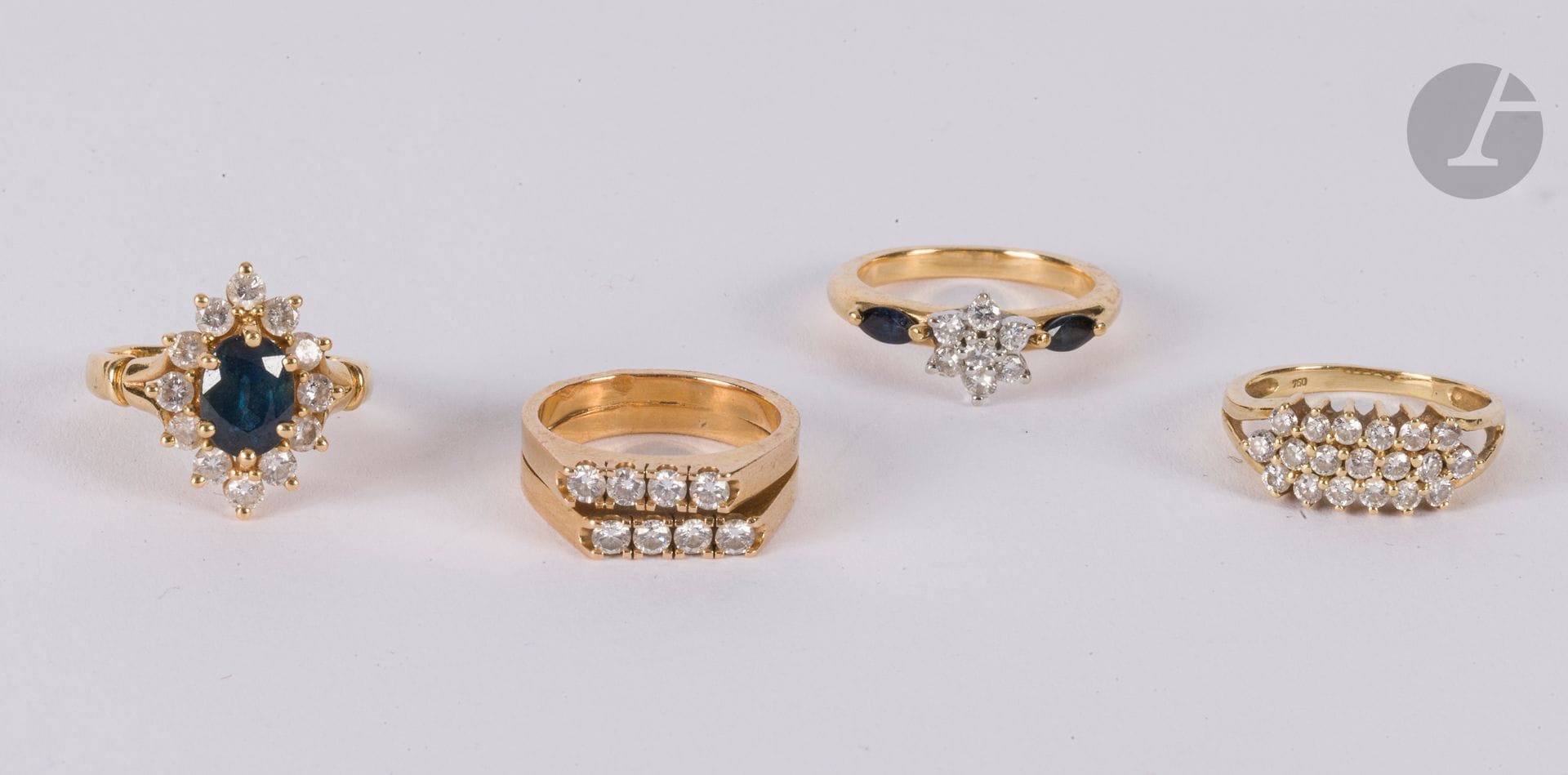 Suite de 4 bagues en or 18K (750) : l'une sertie d'un saphir ovale entouré de diamants ronds de taille brillant, la 2e ornée de diamants épaulés de saphirs, la 3e agrémentée de deux lignes de diamants, la dernière formée de 3 lignes de diamants. Poids brut : 18 g
