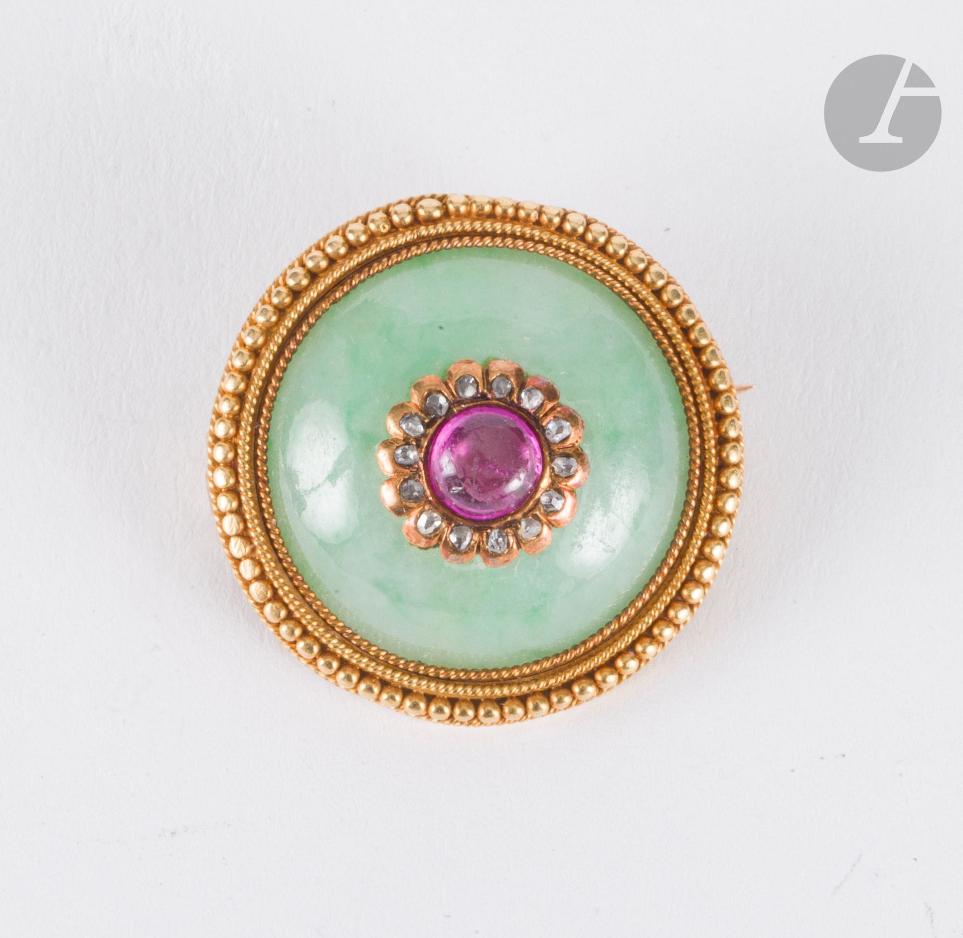 Broche ronde en or 18K (750), ornée d'un jade cabochon centré d'une fleurette en or, le coeur en rubis cabochon, les pétales soulignés de diamants taillés en roses. Diamètre : 2,3 cm environ. Poids brut : 9,3 g (égrisures, choc avec manque de matière)
