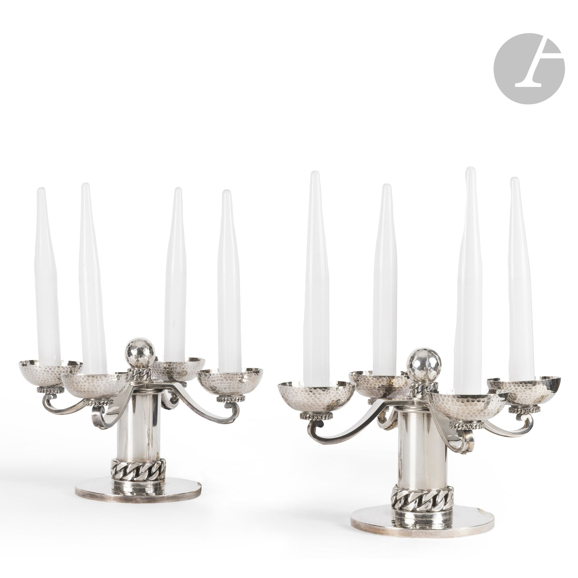 JEAN DESPRÉS (18-1980)
Pair of candelabras, each with four lights, in silver-plated metal - most likely originally electrified.  Circular bases and half-sphere bobèches with hammered surfaces.  The cylindrical shafts are treated smooth and each adorned, on its base, with a large curb chain with flat links. The bobèches are received by small curbets with links. Smooth, moving light arms. Each candelabra topped with a faceted sphere resting on a flat-link bracelet. The perimeter of each base, the ends of the light arms and the bobèches are probably originally drilled to allow electrification. Each candelabra is presented with white opaline candle covers. One bulb cap is damaged and glued back to its base. Unsigned. Without covers: H. 20 cm - L. 33 cm - With covers: H. 39 cm - L. 33 cm