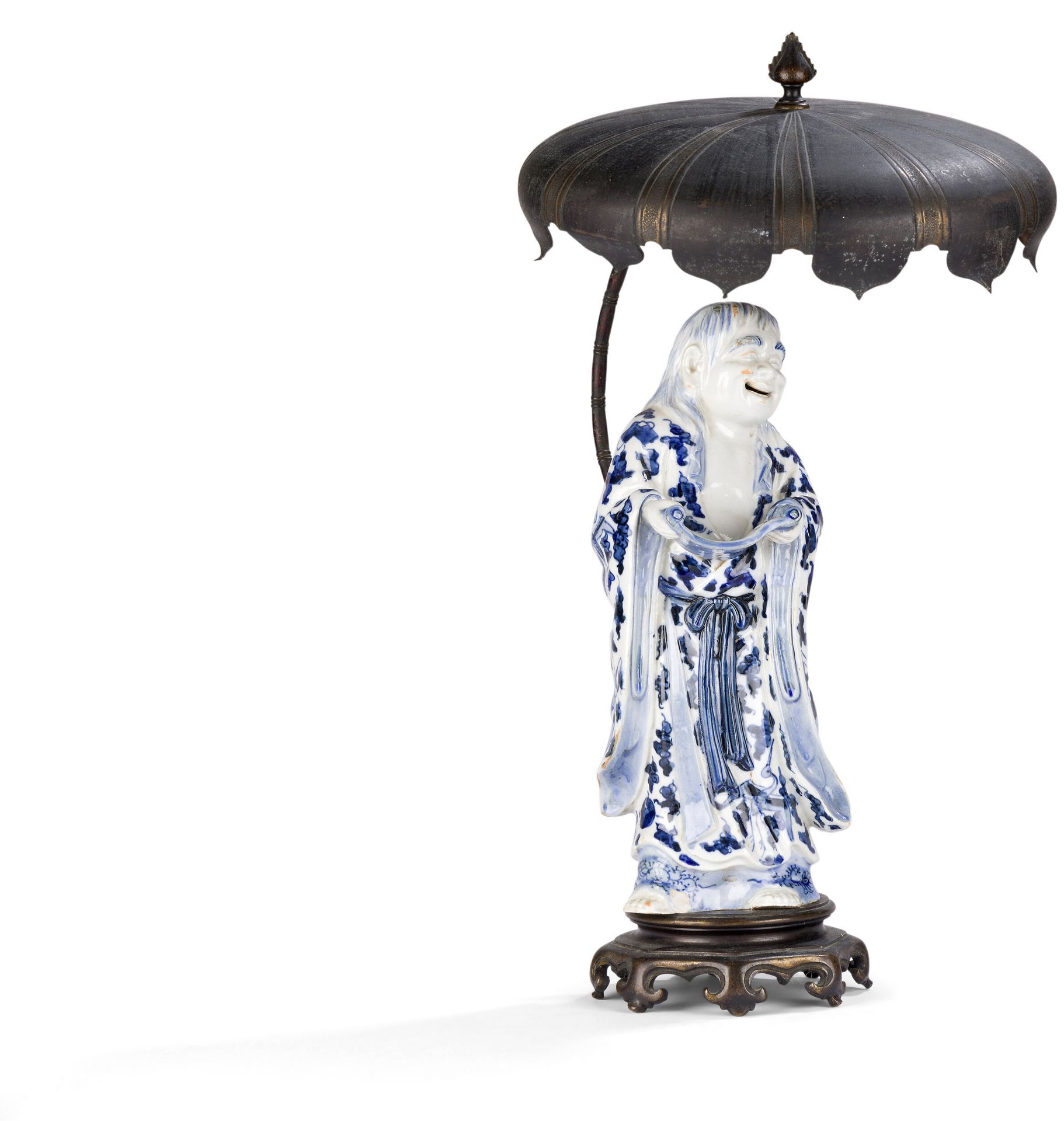 Blue and white porcelain figure of Kanzan, mounted as a lamp, Japan, 19th century
Kanzan, wearing a kimono with blue underglaze enamel decoration featuring tsuru (red-headed cranes), stylized clouds and precious objects, holds a calligraphic scroll and looks at it with a smile. He is mounted on an eight-legged octagonal copper-alloy base, behind which rises a curved stem imitating a bamboo tree, ending in a large parasol.
Height: 28.2 cm
Small chip.

Kanzan, whose name means "cold mountain", was a Zen poet who sought inspiration in nature. Although they look very much alike, he is easily distinguishable from his alter ego Jittoku, as he always holds a scroll of parchment in his hands.