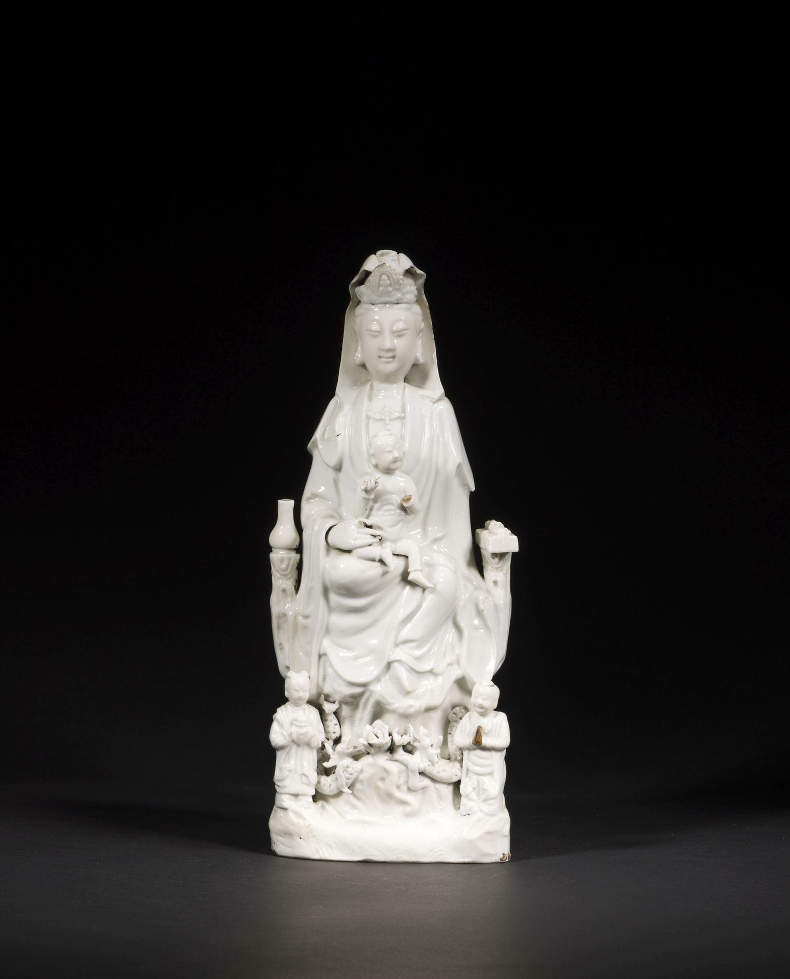 Large Chinese white porcelain Guanyin, China, Dehua, 18th century
Seated with her right leg bent and her left leg dangling in a position known as vamardhaparya?ka, holding a seated child. On one side of her throne, a lustral water bottle on her right, a scroll and sutra books on her left. At his feet, his two acolytes LongNü, holding the Pearl of Light, and ShanCai TonZi, hands in adoration. Between them, on a terrace, a lotus flower surrounded by two facing dragons.
Height: 37 cm
Minor accidents.