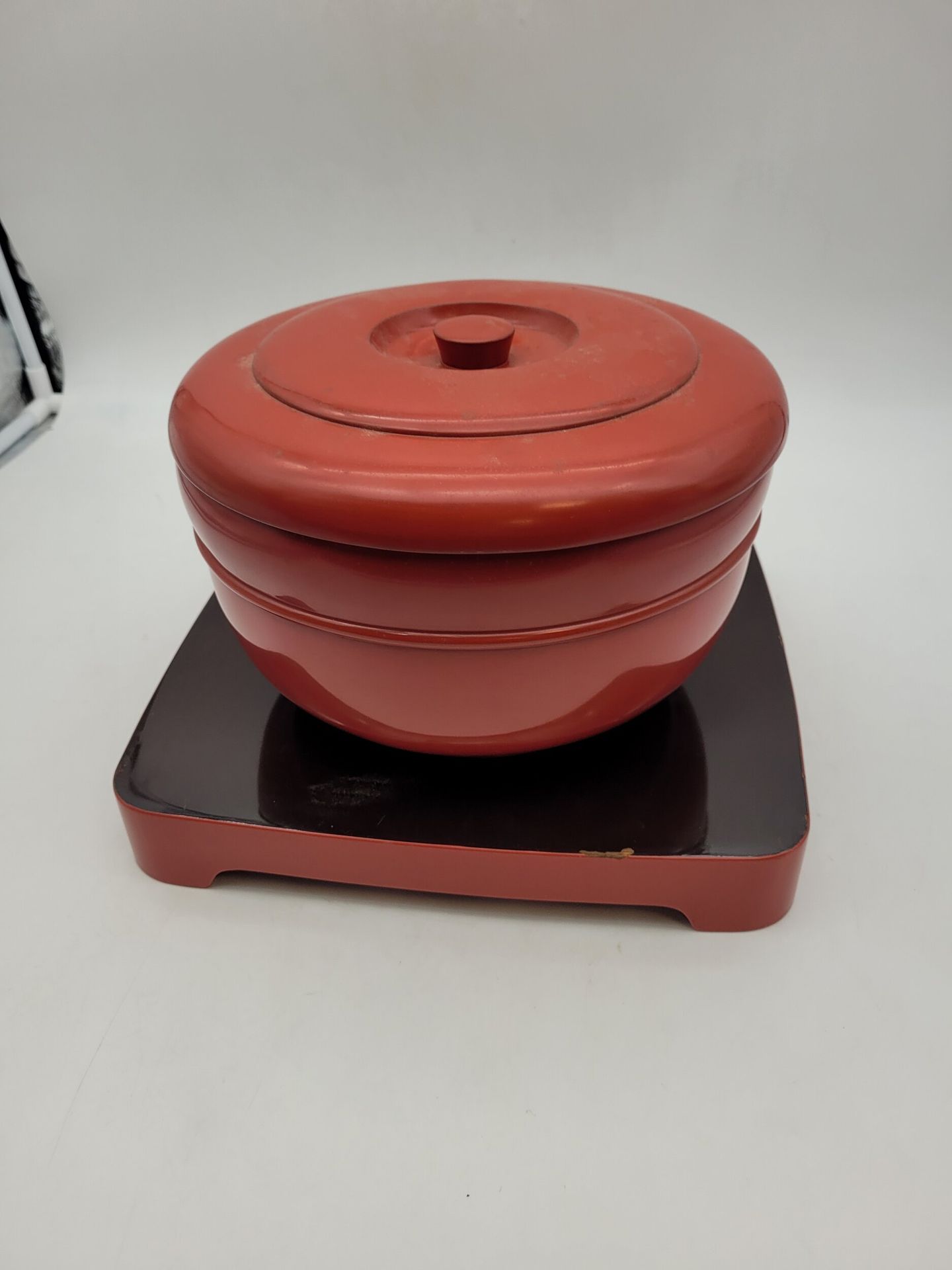 A round lacquered wood box and tray, Japan, Meiji-Taisho period
Cylindrical box of the o hitsu type with a flat grip lid, standing on a small circular foot. Shu nuri (red) lacquered, while the underside is black lacquered. It rests on a square tray with a rounded edge and four feet, the top of which is black lacquered and the rest shu nuri lacquered.
H. box: 16 cm; diam.: 24 cm; tray: 30.3 x 30.3 cm
Chips and scratches.