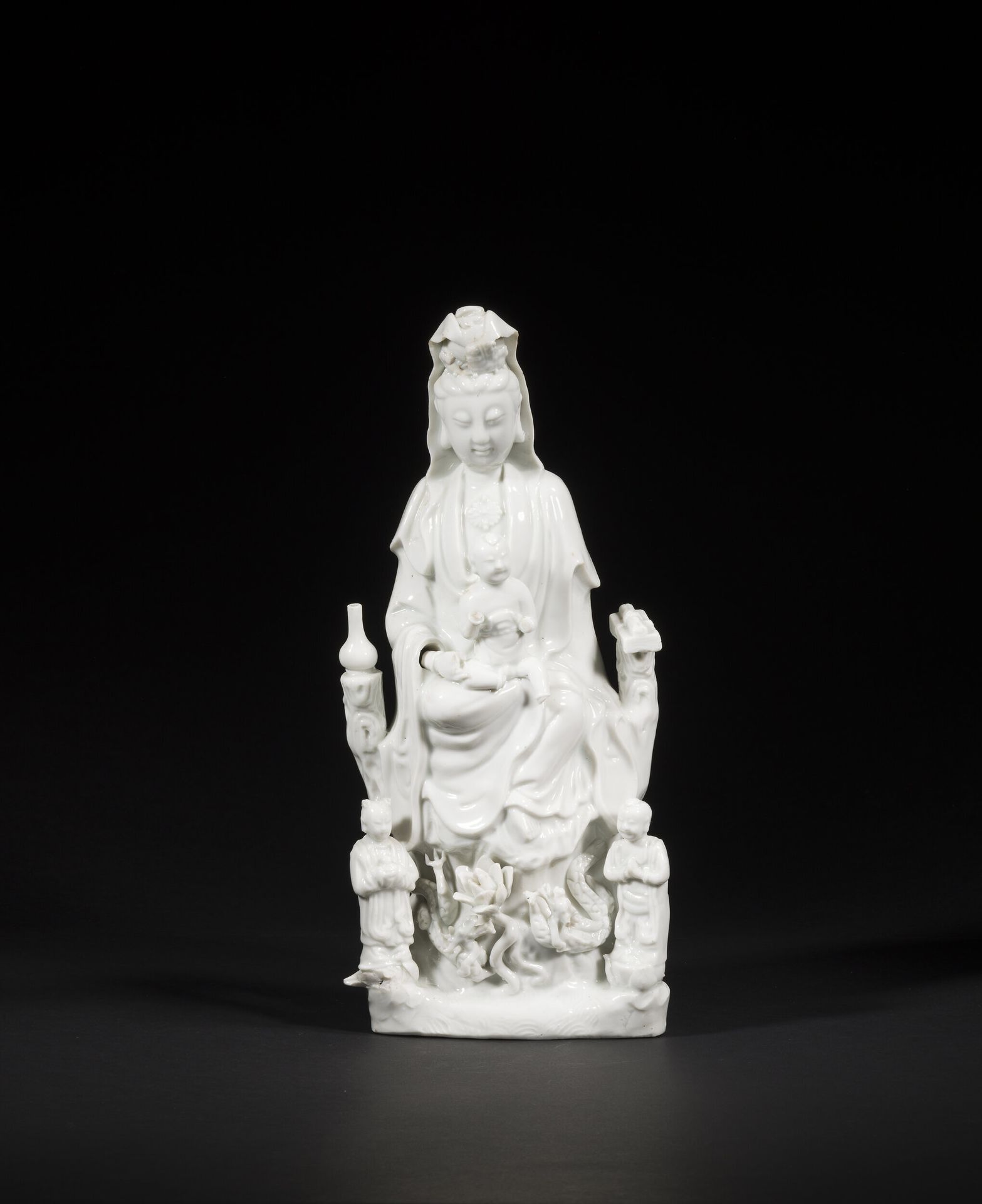 Large Chinese white porcelain Guanyin, China, Dehua, 18th century
Seated, holding a child, with her right leg bent and her left leg dangling in a position known as vamardhaparya?ka. On one side of her throne, a bottle of lustral water on her right, a scroll and sutra books on her left. At his feet, his two acolytes LongNü, holding the Pearl of Light, and ShanCai TonZi, hands in adoration. Between them, on a terrace, a lotus flower surrounded by two facing dragons.
Height: 36.3 cm
Minor accidents.