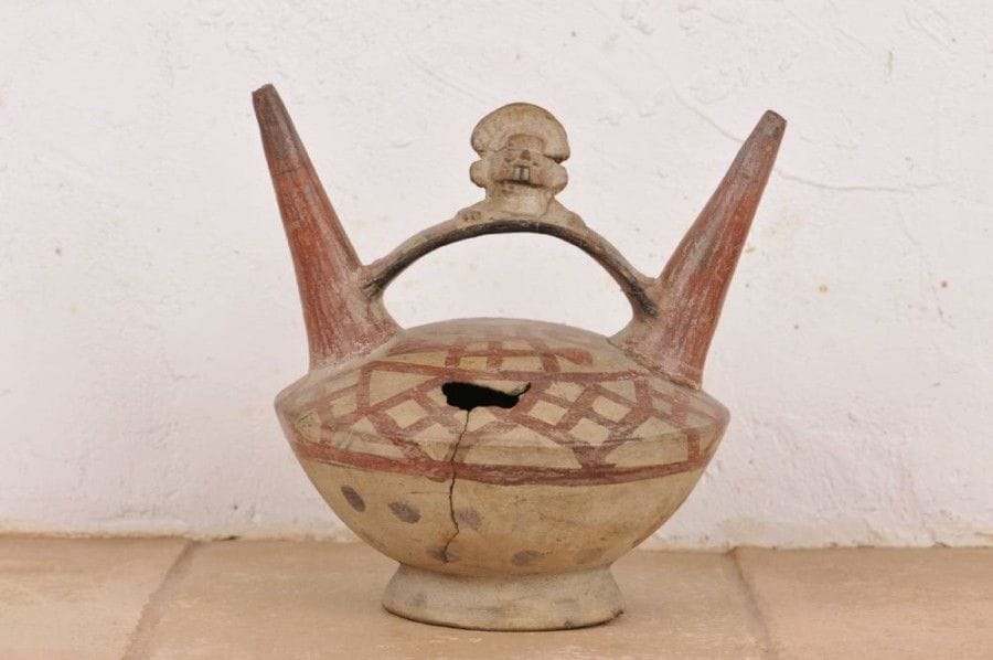 Vase à double goulot décoré d’un personnage sur l’anse médiane. Terre cuite brune à engobe crème. Décor géométrique de couleur rouge sur le haut de la panse qui contraste avec le décor de points noirs dans la partie basse. Culture Chimu - Lambayeque, Pérou, 1100 - 1400 ap. J.-C. H : 19 cm - Diam : 20,5 cm État : cassé et recollé. Deux petites parties manquantes sur le haut de la panse et une en dessous.