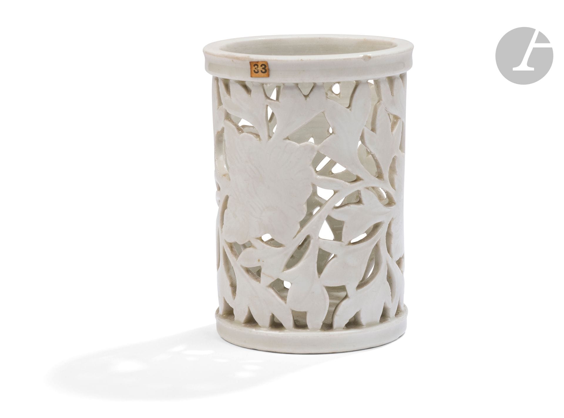 Pot à pinceaux, bitong, Chine, Dehua, époque Kangxi (1662-1722)
Le corps est recouvert d’un émail couleur ivoire, sculpté et ajouré de fleurs de pivoine et de feuillage, avec des décors incisés en surface. Anciennes étiquettes de vente sur le bord et en dessous.
Hauteur : 13 cm
Petit éclat au bord.

La datation de certains groupes de blancs de Chine a été réévaluée ces dernières décennies, notamment grâce aux nombreuses pièces découvertes dans l’épave du Hatcher, datée autour de 1643, et à l’étude de pièces inscrites et datées. Des spécialistes comme John Ayers, ainsi que plusieurs expositions organisées par S. Marchant & Sons, ont établi qu’une grande partie de cette production remonte aux dernières années de la dynastie Ming. Un autre pot à pinceaux au décor ajouré de grecques porte une inscription indiquant qu’il fut réalisé par la famille Lin sous le règne de l’empereur Tianqi (1620-1627). Une autre pièce proposée par S. Marchant & Sons en 2006 est datée de l’été de l’année renzi, soit 1612.
Un pot à pinceaux très proche, conservé dans le Schatzkammer de la Résidence de Munich avec son bassin assorti, est orné de riches montures en argent doré surmontées d’une grenouille en émail, probablement réalisées par des orfèvres parisiens. L’objet comporte également une doublure en verre, le rendant utilisable comme aiguière.