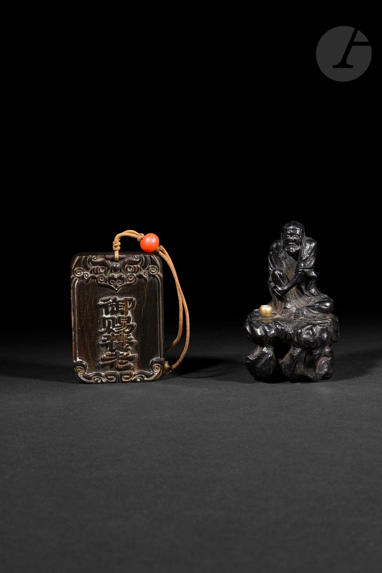 Une plaque en corne et un lohan en bois à patine brune, Chine, XXe siècle
- La plaque est sculptée d’une inscription sur une face et munie d’un cordon avec une bille de verre imitant le corail. 6 x 4,5 cm. - Le lohan se tient assis sur un rocher. H. : 8 cm