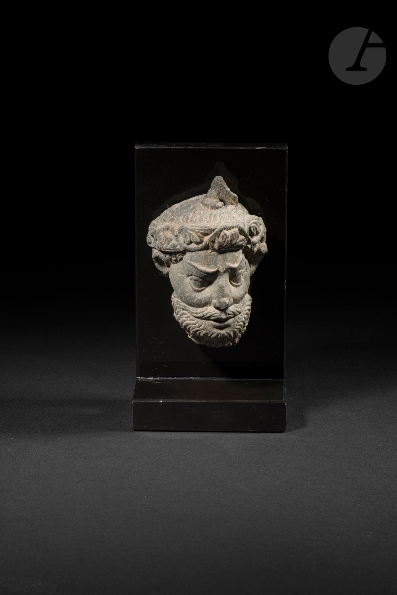 Tête d’homme en schiste gris, art gréco-bouddhique du Gandhara, IIe - IVe siècle
La tête expressive est marquée par des sourcils prononcés au-dessus de grands yeux ouverts, une moustache et une barbe peignées donnant du volume au bas du visage. Le front est ceint au-dessus des oreilles d’un bandeau à motif végétal. Les cheveux sont coiffés et anciennement surmontés d’un chignon.
Hauteur : 10,3 cm
Socle.