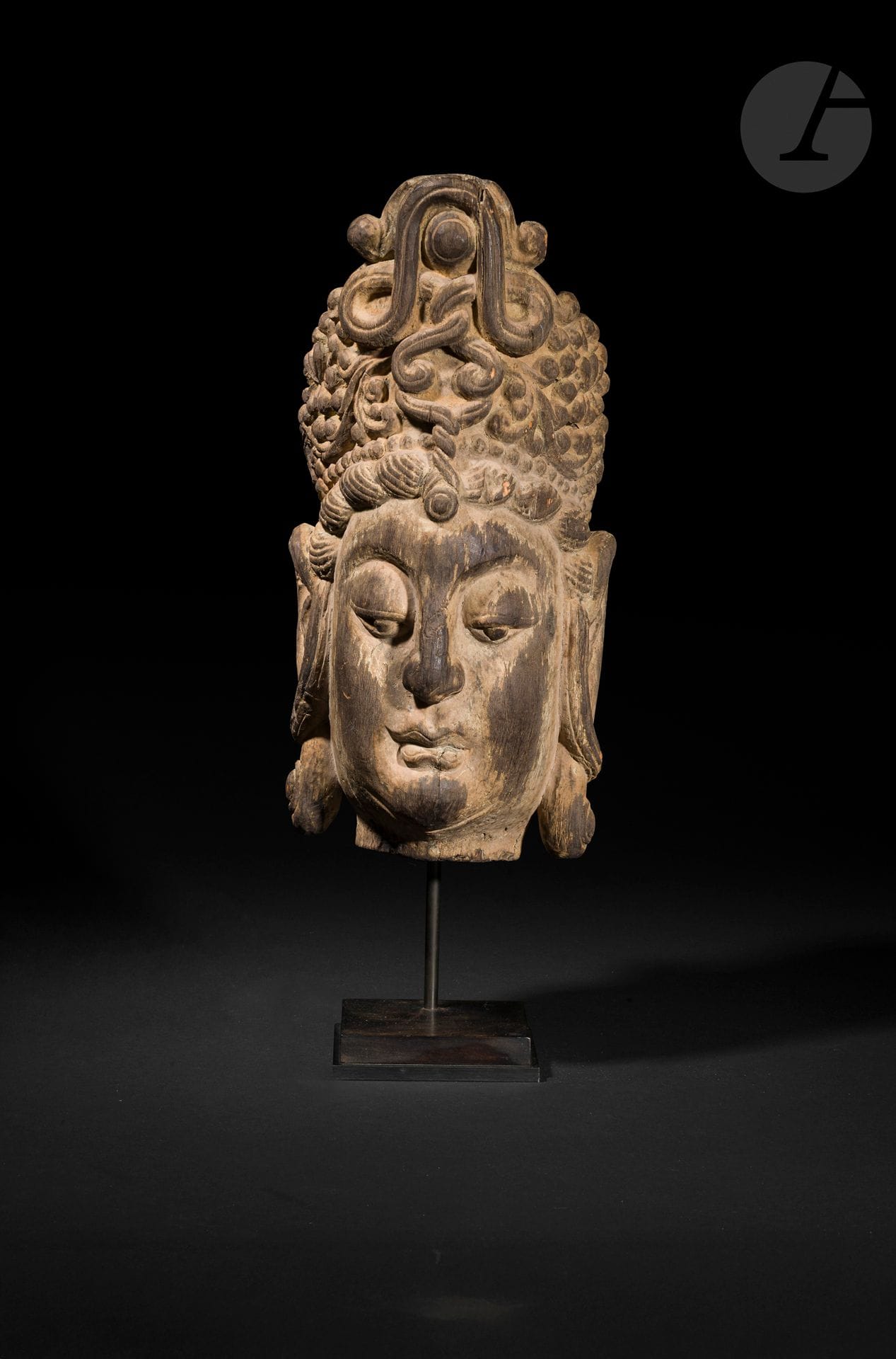 Tête de Guanyin en bois sculpté, Chine, XXe siècle, dans le style des Ming 
Représentation du bodhisattva Guanyin, figuré avec les yeux mi-clos et les traits réguliers. La coiffure est organisée en haut chignon, surmontée d’un diadème orné de motifs sculptés. Les mèches latérales sont traitées en fines ondulations. La surface conserve des vestiges de polychromie, visibles notamment dans les creux de la sculpture. 
Hauteur : 48 cm
Socle.
