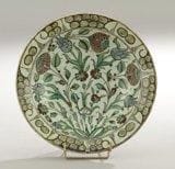Plat tabak au bouquet, Turquie ottomane, Iznik, XVIIe siècle Céramique siliceuse à base annulaire, à décor peint en polychromie sous glaçure transparente sur fond blanc. Bouquet de trois oeillets épanouis, trois tulipes bleues jaillissant d?une touffe de feuillage. Frise de vagues et de rochers stylisés noirs sur le bord. Au revers, alternance de volutes en spirales et nuages tchi. Diam.: 28,7 cm; H.: 6,5 cm An Iznik pottery dish, painted in coloured enamels with flowers, Ottoman Turkey, 17th century.(*)