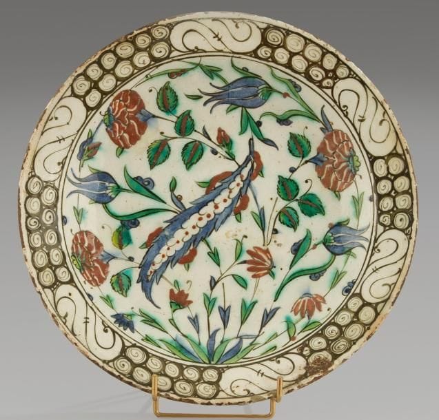 Plat tabak au bouquet tournoyant, Turquie ottomane, Iznik, XVIIe siècle Céramique siliceuse à base annulaire à décor peint en polychromie sur fond blanc sous glaçure transparente. Bouquet tournoyant d?une feuille saz, tulipes et oeillets. Au rebord, frise de vagues et de rochers noirs. Au revers, alternance de volutes en spirales vertes et bleues. D.: 30,5 cm; H.: 5,5 cm An Iznik polychrome pottery dish, decorated with a whirling bunch of flowers, Ottoman Turkey, 17th century.(*)