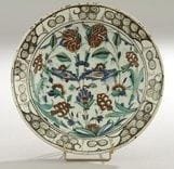 Plat tabak à bouquet symétrique, Turquie ottomane, Iznik, XVIIe siècle Céramique siliceuse à base annulaire à décor peint en polychromie sur fond blanc sous glaçure transparente. Composition symétrique d?un bouquet d?oeillets, et de fleurs composites jaillissant d?une touffe d?herbe. Au rebord, frise de vagues et rochers noirs. Au revers, alternance de volutes en spirales bleues et vertes Diam.: 30 cm et H.: 6 cm An Iznik polychrome pottery dish, decorated with symmetrical flower motifs, Ottoman Turkey, 17th century.(*)