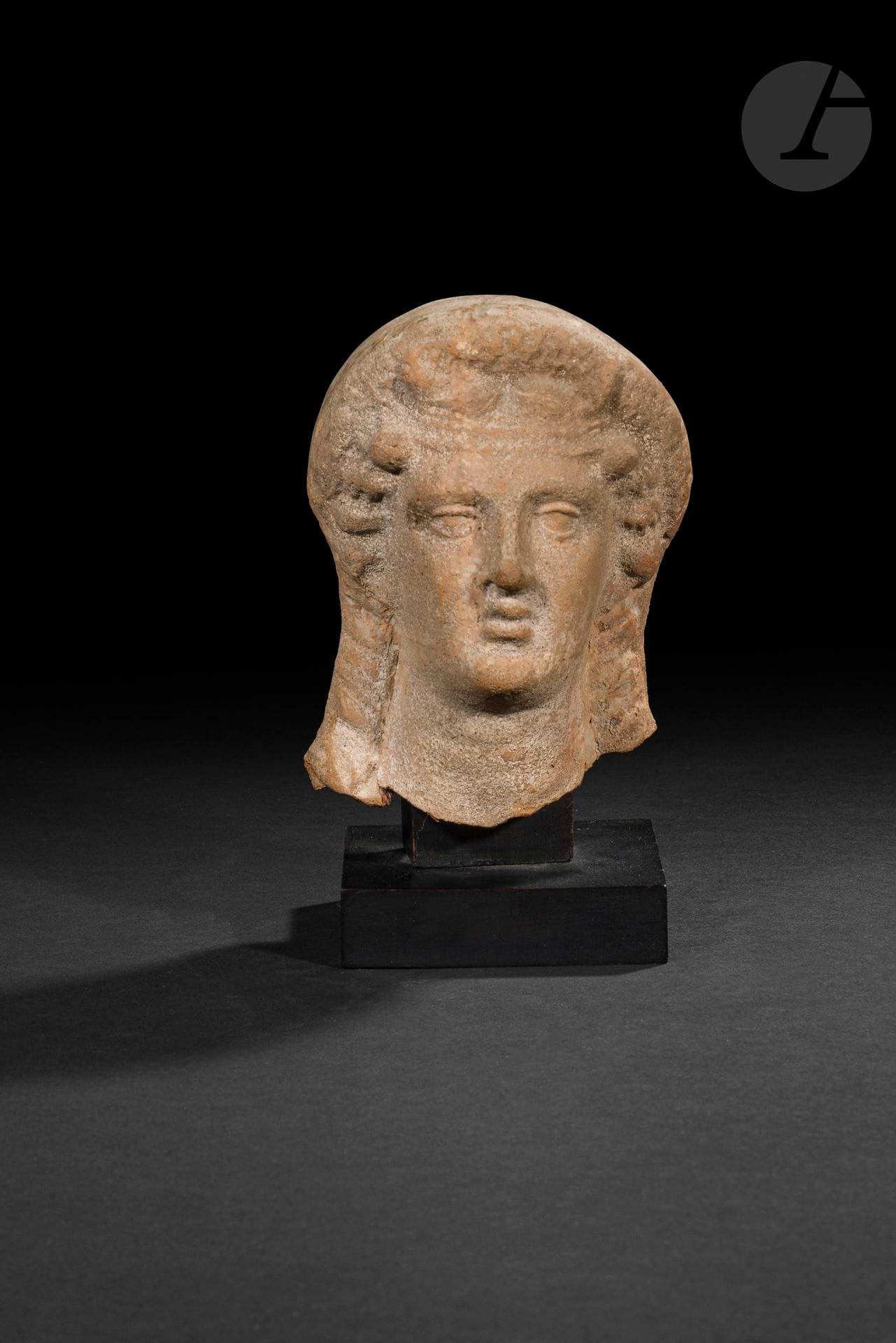 Tête féminine à la coiffure retenue par un bandeau et une couronne
Deux boucles encadrent son visage. 
Terre cuite. Cassée-collée.
Art étrusque, IV-IIIe siècle avant J.-C. 
Hauteur : 13 cm