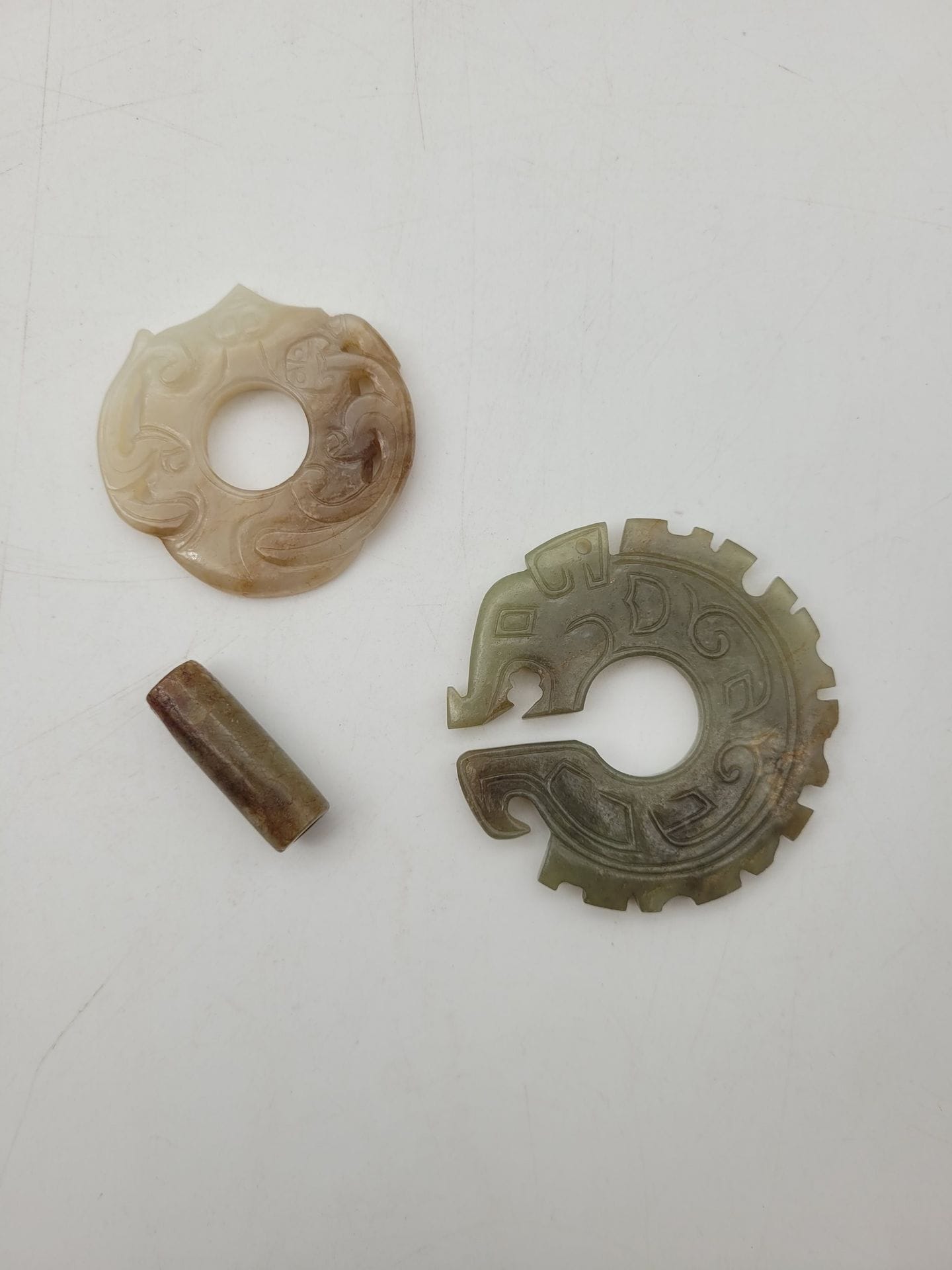 Two archaic-style jade ornaments, China, 20th century
- One in green jade in the form of a stylized dragon coiled around itself. D: 5.7 cm; - the other in celadon and rust jade carved with a chimera. D: 4.5 cm.
An oblong jade bead is attached.
