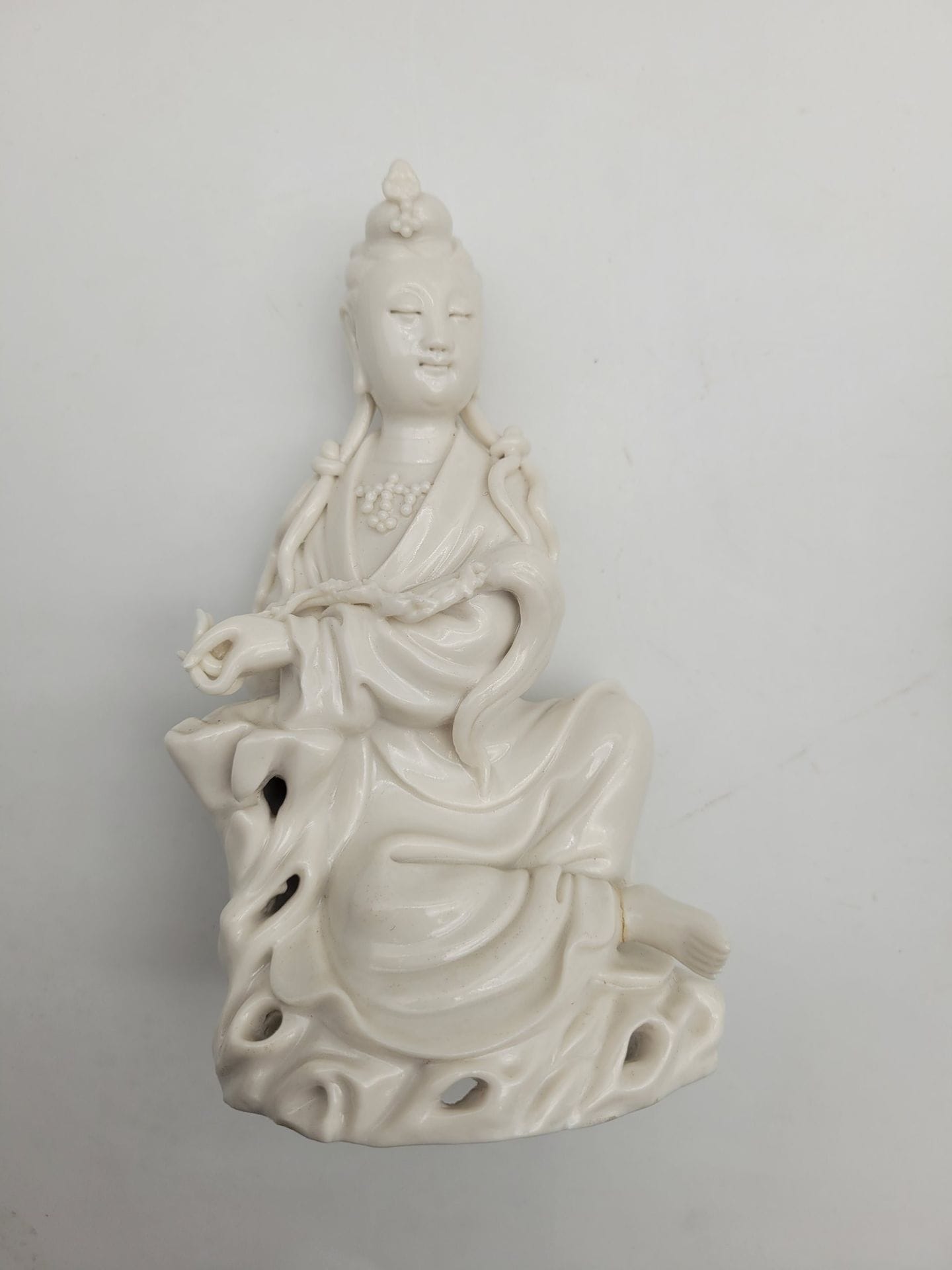 Small Chinese white porcelain guanyin, China, Dehua, 19th - 20th century
She is depicted seated on a pierced rock, wearing a long pleated robe and a pearl necklace. Her serene face features a slight smile, the forehead is adorned with the urna and the earlobes are elongated. The hair is gathered into a small bun, adorned at the front with an ornament, and divided at the back into two long locks passing over the front of the shoulders and knotted to form two new locks. His left arm rests on a rock, and his hand holds a fuzi. The naked right foot protrudes from the bottom of the dress. On the back is a molded double-gourd mark, difficult to identify.
Height: 13.5 cm
Foot and top of headdress ornament reattached.