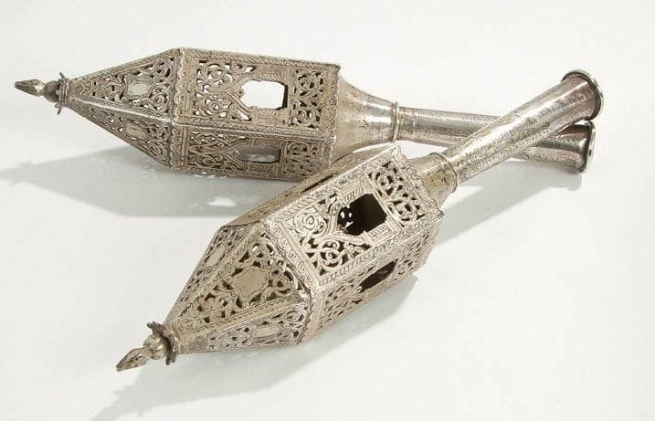 PAIRE DE RIMONIM en argent gravé et ajouré. Ils portent tous deux sur la tige une dédicace en hébreu: «offerts par les familles Zenou et Sebban en 1926». L'un des deux porte en hébreu l'inscription: «oeuvre de Nissim Partouche», l'autre: «oeuvre de Lalou Partouche». La famille Partouche était une famille d'orfèvres réputée à Alger. Comme souvent les grelots manquent. Ancien travail d'Afrique du Nord. Longueur: 30 cm - Poids des deux: 534 g