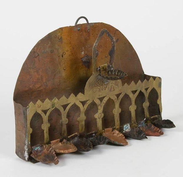 LAMPE DE HANNOUCA en cuivre et en laiton. Dosseret en cuivre, partie avant en laiton ajourée de mihrabs sur laquelle viennent se fixer les godets. Complète de tous ses godets ainsi que de son shamash. Afrique du Nord (probablement Algérie), XIXe siècle. Hauteur: 18 cm - Longueur: 25 cm Bel exemplaire dans son jus.