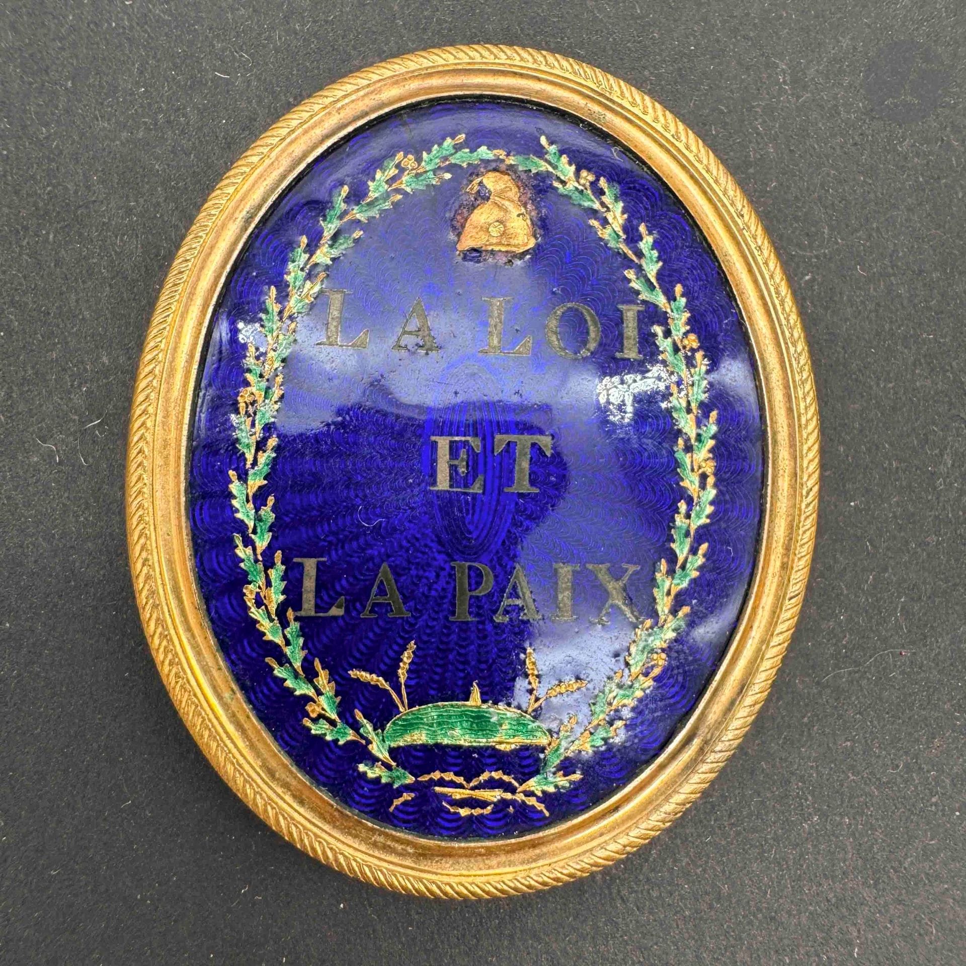 FRANCE 
Insigne de juge de paix. 
En laiton doré, à centre émaillé à décor de couronne de feuilles de chêne, bonnet phrygien (manque) et la mention «la loi et la paix». Revers en métal blanc à quatre anneaux d’attache. 46 x 36 mm
A.B.E. Époque révolutionnaire.