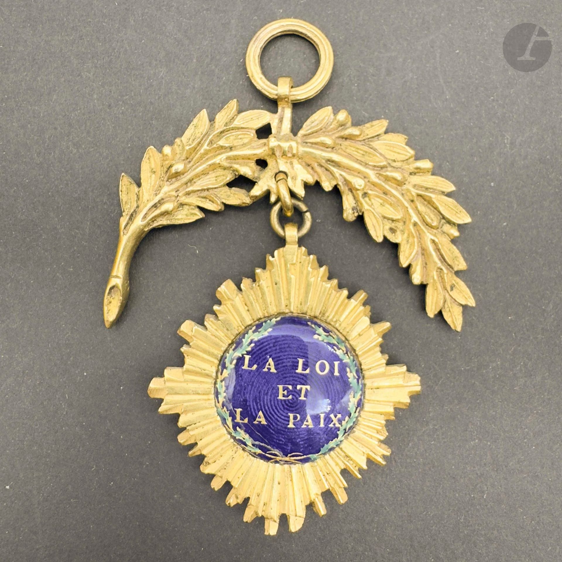 FRANCE
Insigne de juge de paix en bronze doré composé d’une étoile rayonnante à quatre pointes. Centres en émail bleu marqués « la loi et la paix » dans des couronnes de feuilles de chêne. L’étoile surmontée d’un motif à la branche de laurier. Anneau cannelé. 
69 x 41 mm 
A.B.E. (griffures à un centre et traces de réparation à l’étain). Époque révolutionnaire.