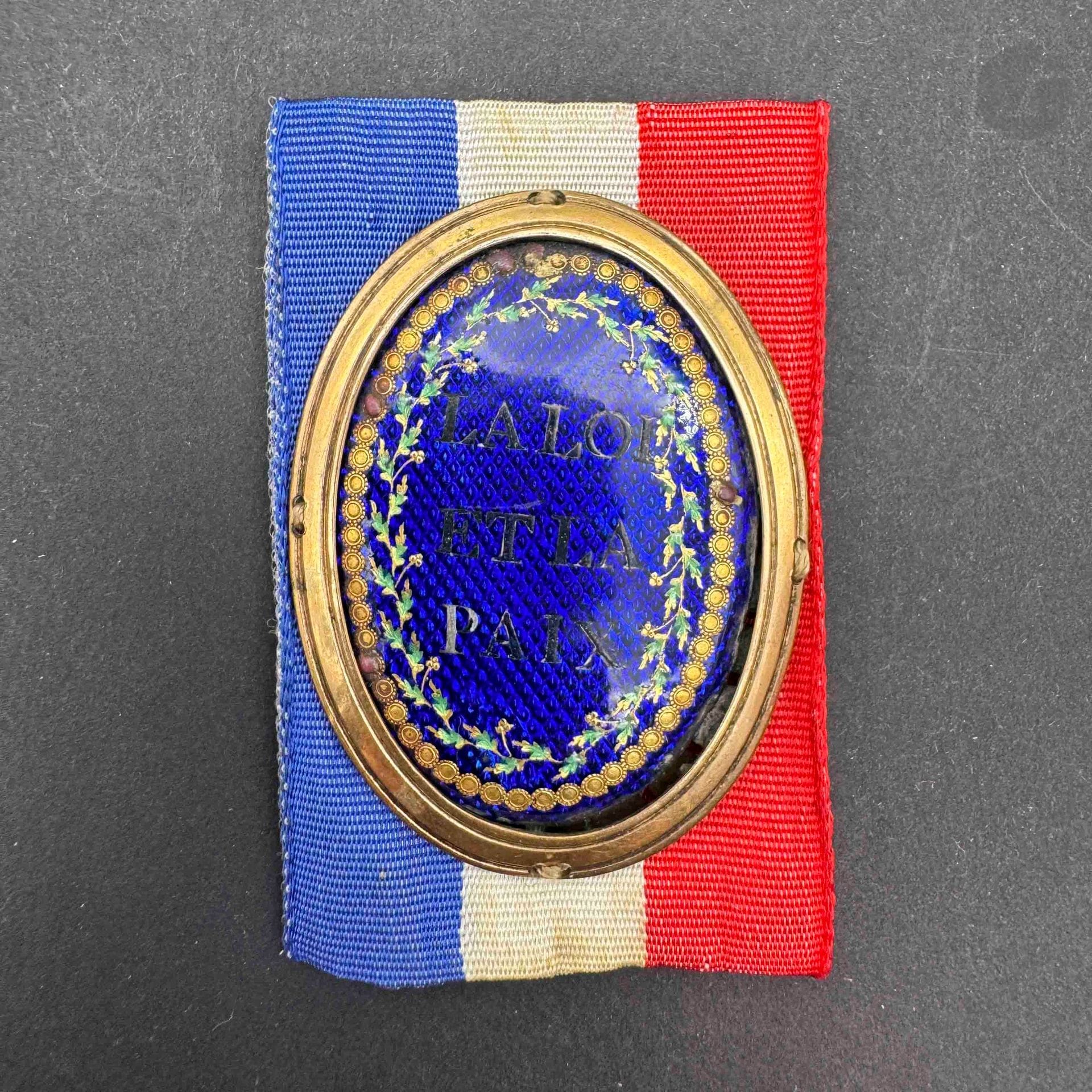 FRANCE
Médaillon de juge de paix en laiton doré à centre émaillé bleu sur fond de croisillons marqués à l’argent « la loi et la paix » dans un entourage de feuilles de chêne. 
Présenté postérieurement sur un ruban tricolore. 
60 x 40 mm 
A.B.E. (petites usures). Époque révolutionnaire.