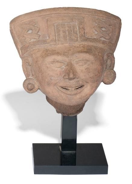 Tête de souriante. Joli visage souriant laissant apparaître les dents du devant. Les yeux en amande sont rapprochés selon les critères de beauté de l?art du Veracruz. Une coiffe en bandeau ornée de motifs symboliques ainsi que des boucles d?oreilles circulaires agrémentent l?ensemble. Trou de cuisson de chaque côté de la coiffe. Terre cuite brune. Culture Veracruz, Mexique 450 - 750 ap. J.-C. H: 15 cm - l: 15 cm - Ep: 9 cm Provenance: collection privée, Paris