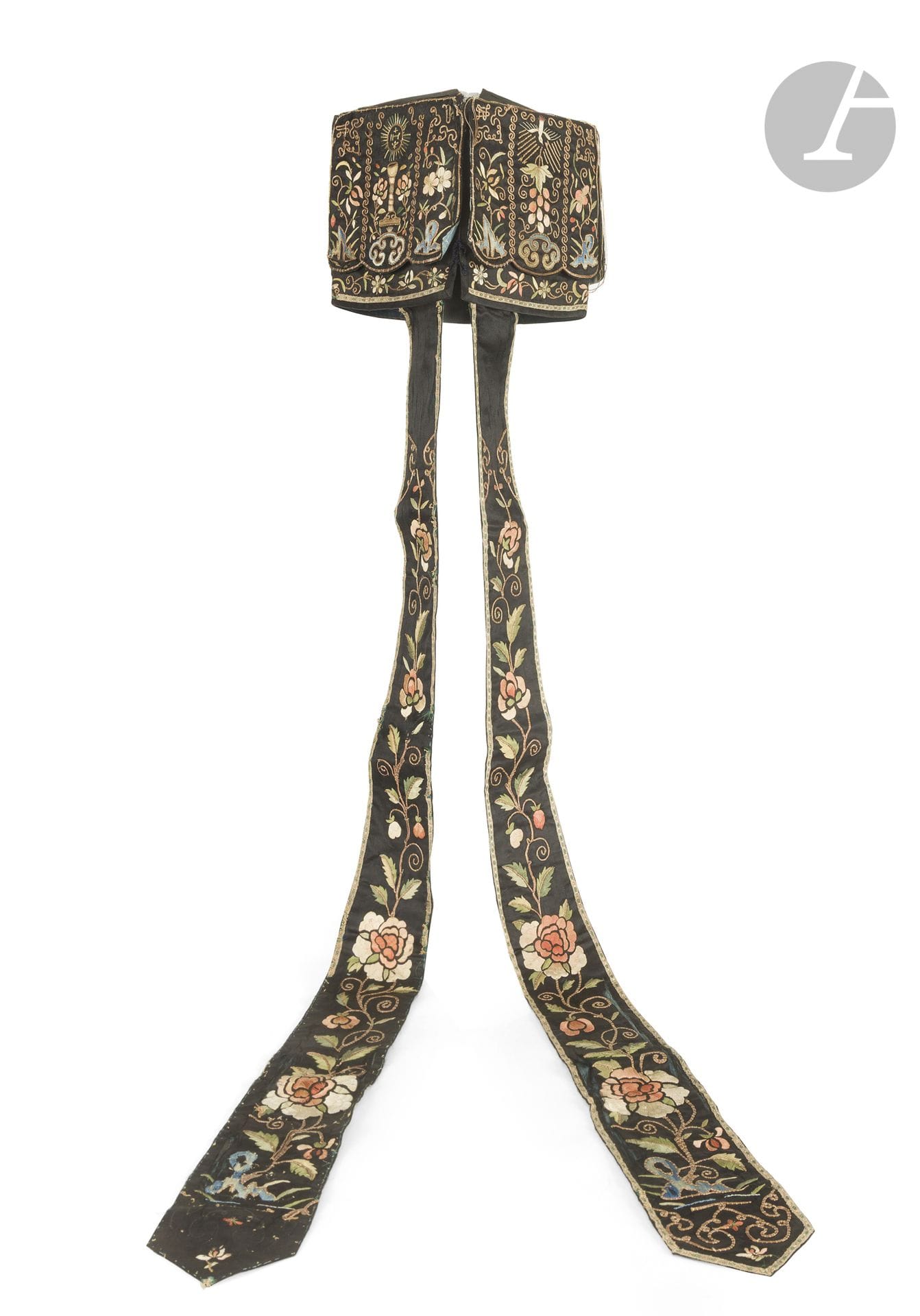 A Jijin liturgical headdress—祭巾—worn by Catholic priests and missionaries in China in the late 19th and early 20th centuries
Made of black silk satin, square in shape and foldable flat for storage and transport, embroidered with multicolored silk threads and gold metallic thread depicting Christian symbols on each side: the Chalice and the radiant Host bearing the coat of arms of the Society of Jesus, accompanied by a cross and the three nails of the crucifixion. The Dove of the Holy Spirit flying in a halo of light above a stylized bunch of grapes symbolizing the blood of Christ. The Sacred Heart of Jesus above a crescent moon surmounted by red flames and three flowers. It represents divine love and devotion to the Sacred Heart, which was widespread among Jesuit and Lazarist missionaries. The Immaculate Heart of Mary (or second Heart), surmounted by a four-petaled flower symbolizing the cross, with the three nails of the Passion depicted at the bottom. These motifs are framed by Chinese-inspired borders depicting ruyi heads, plum blossoms and orchids, lotus flowers, rocks, and endless knots. The headdress is extended by two long ribbons embroidered with peony flowers.
16 x 16 cm; length: 102.5 cm

The jijin—祭巾, literally “ceremonial cloth”—is a liturgical headdress adopted by Catholic priests and missionaries in China beginning in the 17th century. Its use was officially authorized in 1615 by Pope Paul V, at the request of the Jesuits, in order to accommodate the customs and conventions of Chinese society. Indeed, appearing bareheaded before a person of higher rank was traditionally considered a sign of disrespect or humiliation. The jijin thus allowed the priest to remain covered during the celebration of Mass, while affirming his religious status. Its square shape, inspired by traditional Chinese headdresses, reflects the missionaries’ adaptation to local customs. Usually made of brocaded silk, it consists of a square cap with panels extending down around the head and down the back. The corners are often adorned with cords or ties. The ornamentation, deeply Christian in nature, thus combines a Chinese form with symbols of the Passion of Christ, the Crucifixion, and the Catholic faith. The use of the jijin was officially approved by Clement X in 1673, and it remained part of the Chinese Catholic clergy’s attire for several centuries. It thus stands as a remarkable testament to the convergence of Chinese sartorial tradition and Catholic liturgy. Its use gradually declined in the early 20th century with the Westernization of ecclesiastical practices and virtually disappeared after the First Chinese Council in Shanghai in 1924. The surviving examples are rare today and offer a particularly valuable insight into the history of the early Catholic missions in China.