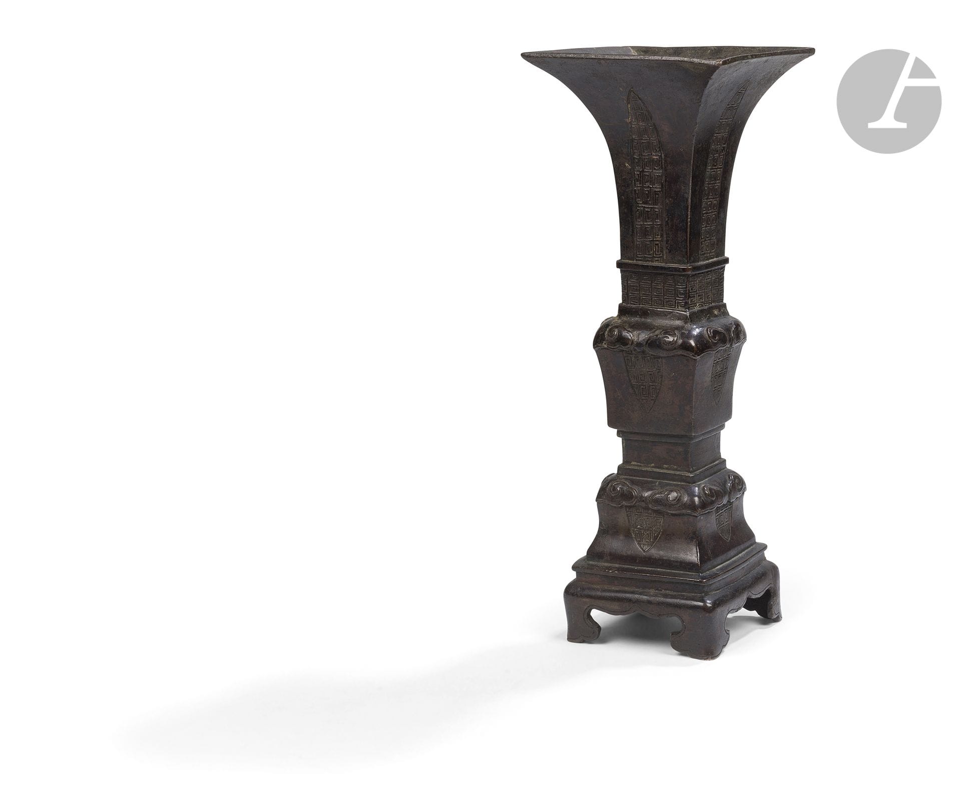 Vase fanghu archaïsant en bronze, Chine, époque Ming, XVIe siècle 
De section quadrangulaire, reposant sur un pied quadripode et présentant un haut col largement évasé, le vase est rythmé par plusieurs registres séparés par des bandeaux en léger relief. Les différentes faces sont ornées de réserves géométriques encadrant des décors de grecques, tandis que les angles des registres sont soulignés de motifs de nuages stylisés en forme de lingzhi - 靈芝. La composition, organisée selon une alternance de panneaux et de bandeaux décoratifs, reprend de manière stylisée le vocabulaire des bronzes archaïques chinois. La surface présente une patine brune sombre et homogène. 
Hauteur : 38,2 cm
Usures et petites irrégularités de surface, réparation au fond.