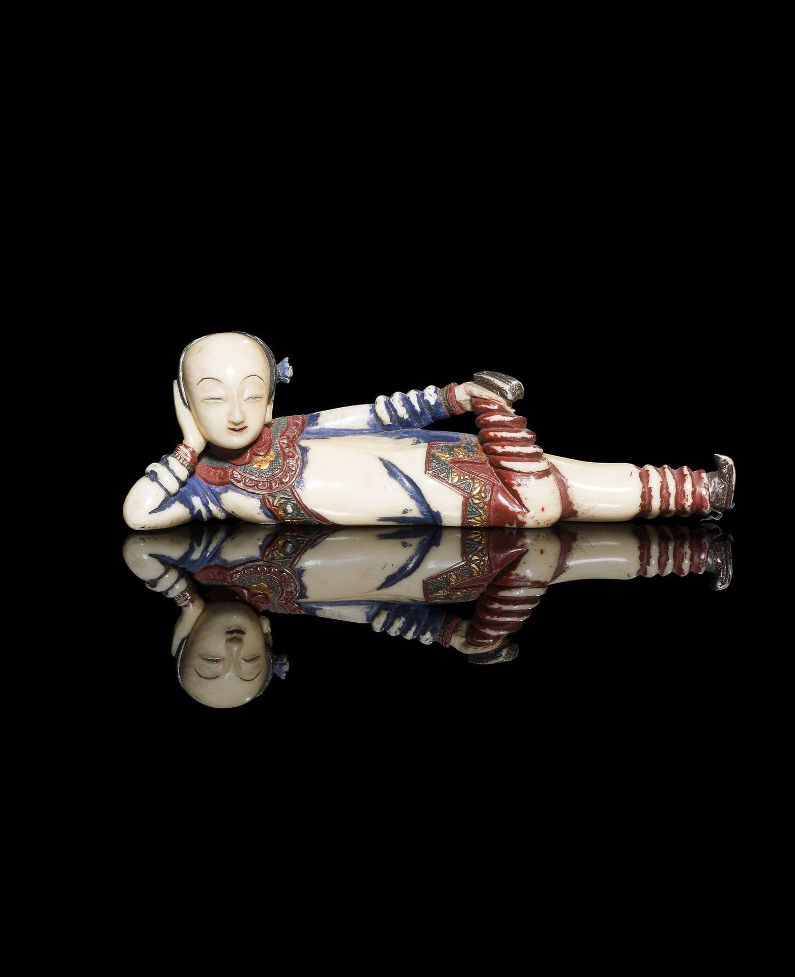 Tabatière en ivoire sculpté en forme d’une femme allongée, Japon, époque Meiji (1868-1912) - Taisho (1912-1926)
Le visage nous fait face et la joue repose contre la paume de la main droite dont le bras posé au sol est replié. La jambe gauche est tendue alors que la droite, pliée, remonte de façon perpendiculaire vers la main du bras gauche tenant le pied. Le visage est doux et souriant, les cheveux plaqués sont ornés d’une petite fleur. Elle porte une tunique et un pantalon anciennement rouge et bleu, bordés de passementeries à décor de fleurs avec des traces de dorure. Le pied de la jambe gauche est amovible, formant le bouchon du flacon. Restes de polychromie et dorure.
Longueur : 11,4 cm ; poids : 37 g