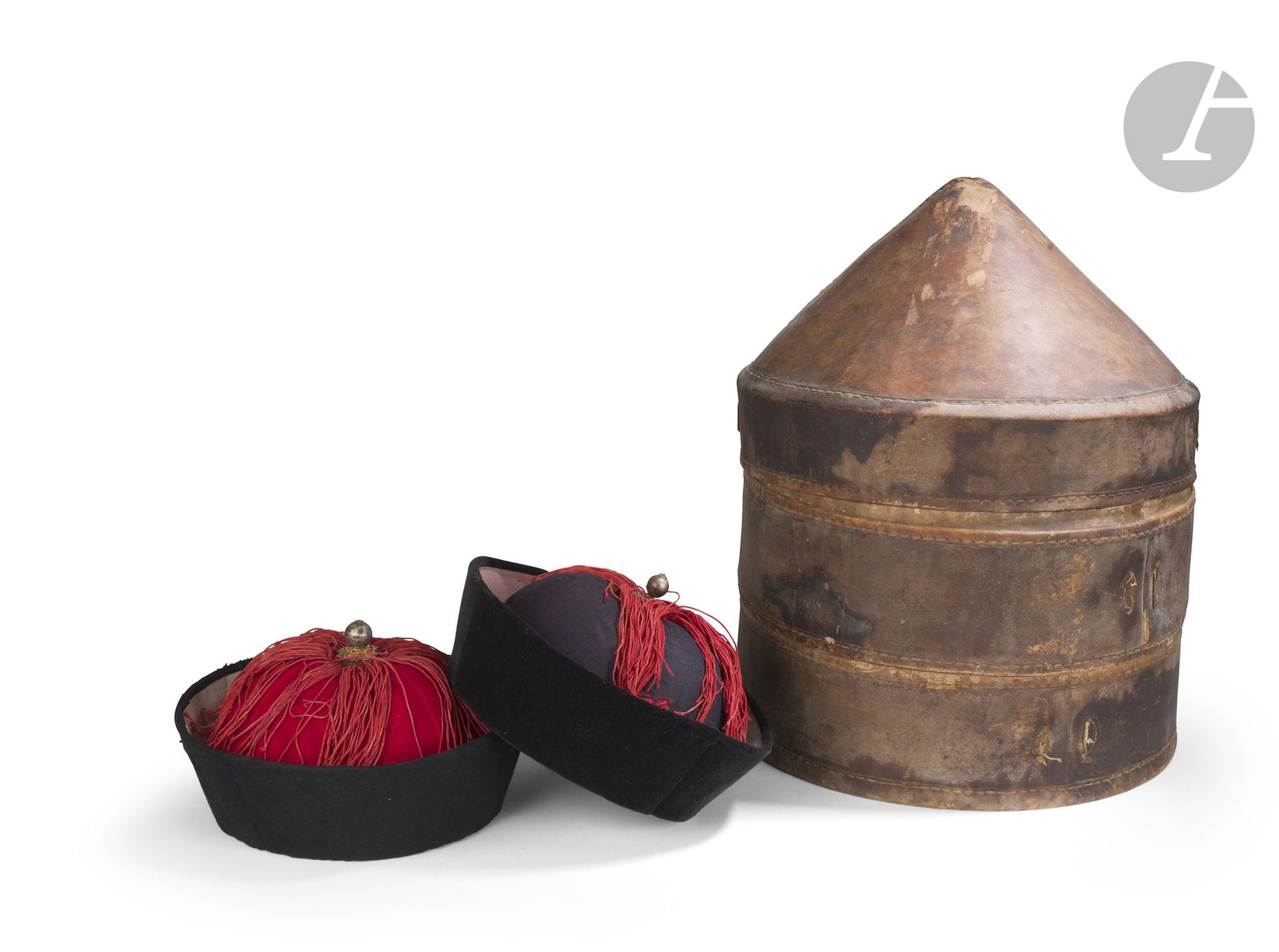 Deux guanmao, chapeau d’hiver d’officiel mandchou, dans leur boîte de transport, fin du XIXe siècle
A bord relevé, l’un est recouvert de soie damassée à décor de bambous et fleurs de prunier, l’autre est en velours noir. La calotte est agrémentée d’une frange de fils de soie rouge hóngyīng rayonnant depuis le sommet où est fixé un bouton en alliage de cuivre indiquant le 9e rang dans la hiérarchie mandarinale. Ils sont conservés dans une boîte de transport en cuir de forme cylindrique avec un couvercle conique. L’intérieur montre deux étages.
Hauteur : 45 cm ; diamètre : 36 cm
Usures et manques.

A l’intérieur du couvercle, une étiquette imprimée en noir sur papier, portant le nom de la maison Dafengtai - 大豐泰, une adresse au marché aux chevaux de Linqing (Shandong), la mention d’une fabrication de coffres et boîtes en cuir, ainsi qu’un gage de qualité pour la clientèle.