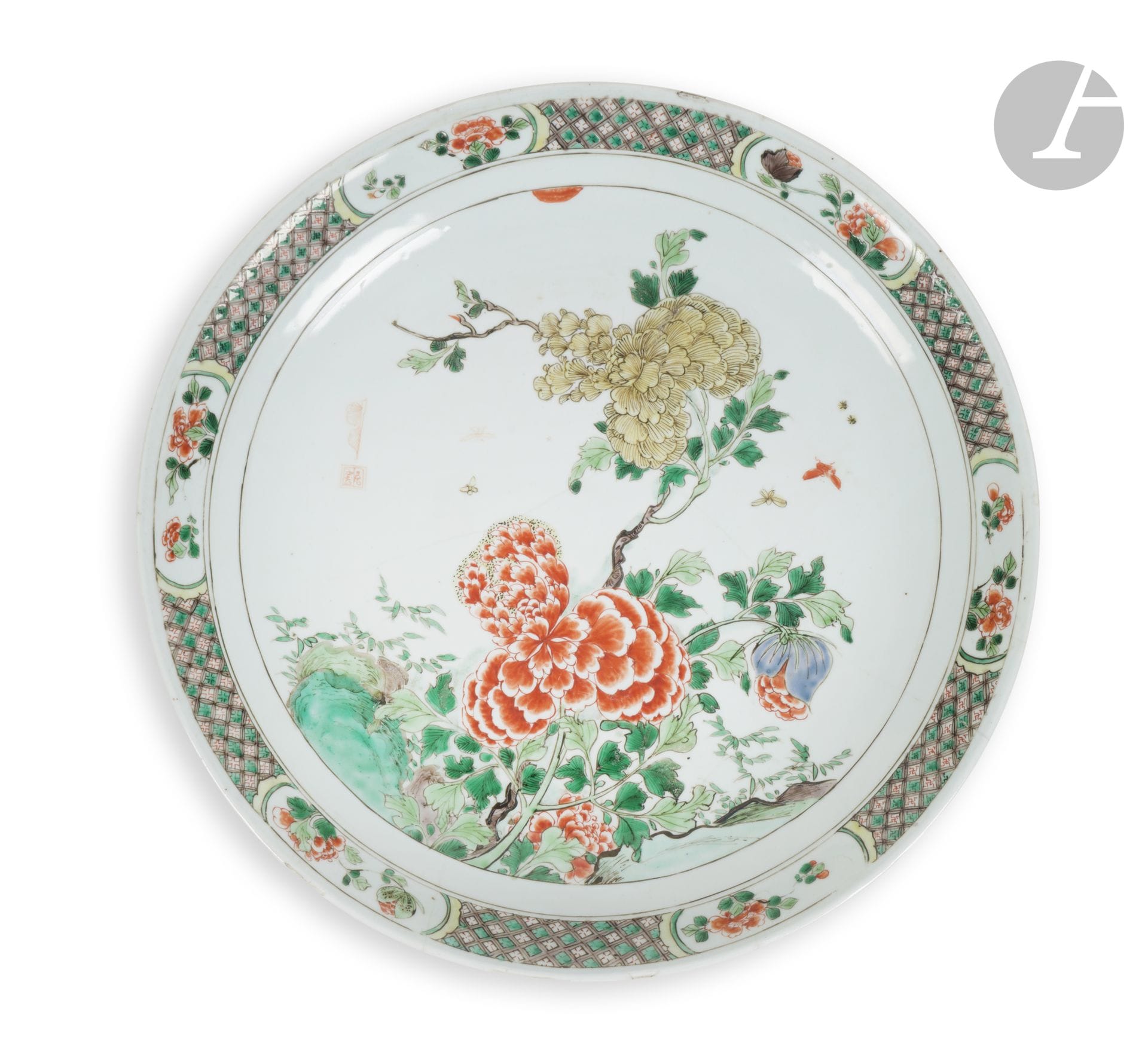 Plat en porcelaine de la famille verte, Chine, époque Kangxi (1662-1722)
À décor central de grandes pivoines et leurs feuillages s’élevant au-dessus de rochers et bambous autour desquelles volent des papillons et petits insectes. Sur la gauche, une marque fantaisiste dans un carré accompagnée d’une feuille. Le marli est décoré de six cartouches à décor de pivoines sur un fond de frises géométriques, fleurettes et svastika dans des losanges. Au revers, une marque au champignon émaillée en bleu sous couverte dans un double cercle et la marque d’inventaire incisée « N: 131 工 » de la collection d’Auguste II le Fort, électeur de Saxe, roi de Pologne.
Diamètre : 36 cm
Fels et égrenures.

Le marquage « N: 131 工 » correspond à la série de douze assiettes à décor de la famille verte de fleurs, certaines étant également ornées d’un oiseau, documentée dans les inventaires de la Porzellansammlung de Dresde (Staatliche Kunstsammlungen Dresden, inventaire n°324, 1721, Famille verte, p. 830 : « 131. 12 Stk. runde Schaalen, darein allerhand Blumen, in etl. aber ein Vogel gemahlet, 3 Z. tief u. 15 Z. indiam. » – « 131. 12 plats ronds, richement peints à l’intérieur de fleurs, avec, sur certains, un oiseau peint, de 3 pouces de profondeur et de 15 pouces de diamètre »). Deux autres plats de la même série et portant le même numéro d’inventaire incisé sous la base sont conservés au Porzellansammlung à Dresde (PO 6929 et PO 6930). L’entrée correspondante de l’inventaire de 1779 apporte également des précisions sur les circonstances dans lesquelles ces plats furent acquis pour les collections d’Auguste le Fort : « Eod. ao. mense Julii haben Ihro Königl. Mayt. von dem Herrn Geh. Rath von Rechenberg erkauft: » - « En juillet de cette même année, Sa Majesté Royale acheta les articles suivants au conseiller privé von Rechenberg ».