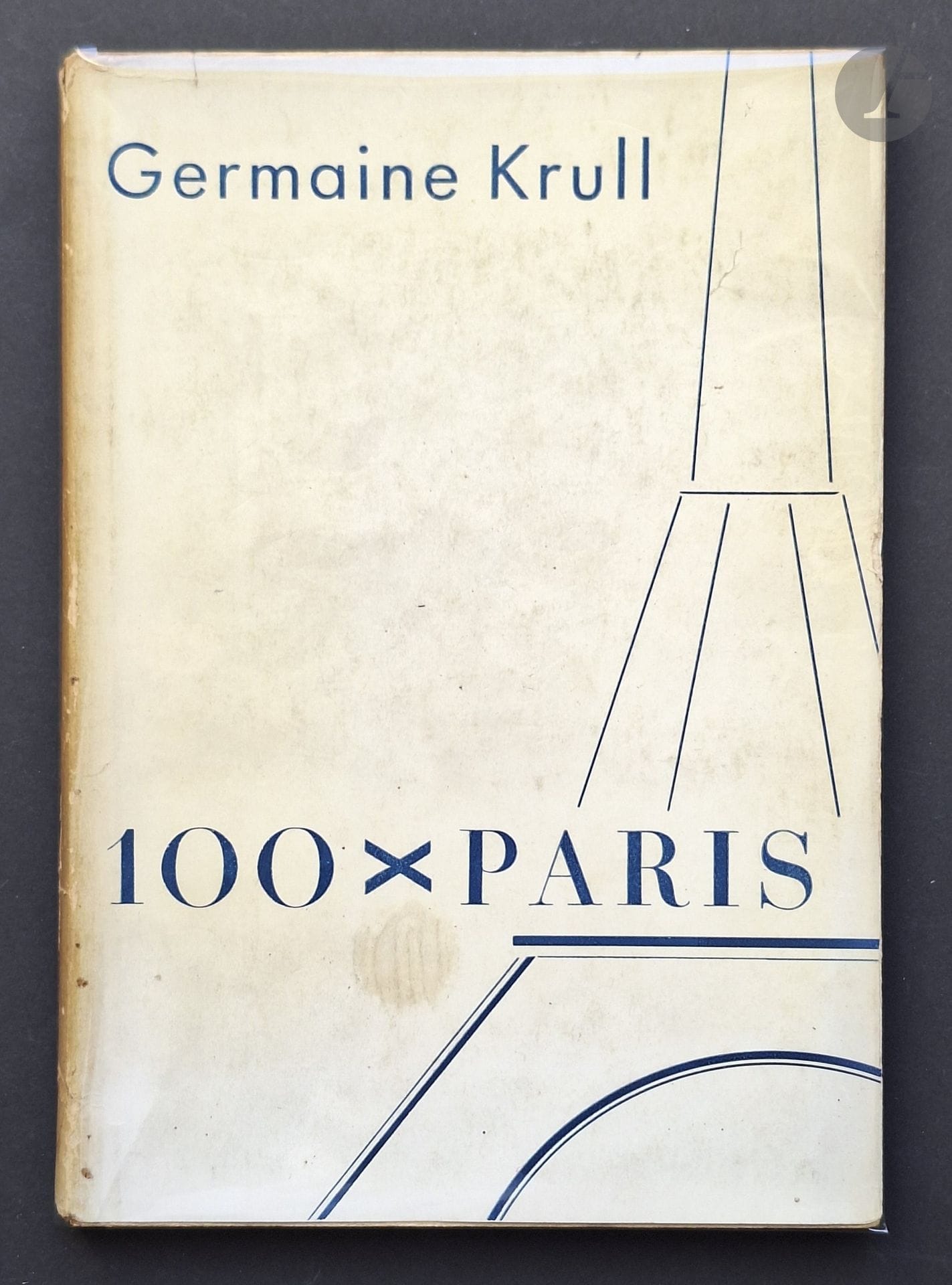 KRULL, GERMAINE (1897-1985)
100 x Paris.
Verlag der Reihe, Berlin, 1929.
In-8 (25 x 18 cm). Édition originale. La série des cents. 31 pp. de texte trilingue (allemand, français et anglais) et 100 héliogravures. Ouvrage broché, sous jaquette illustrée. Plats de la jaquette entiers, mais sans les rabats; dos bruni. Quelques défauts sur les deux premières et deux dernières pages. Rare avec sa jaquette.
Fascinée par Paris comme nombre de ses contemporains photographes, Germaine Krull propose dans 100 x Paris,  une vision inédite de la capitale des années 30. Son regard se pose à la fois sur le quotidien de la ville et de ses habitants. Son oeil particulier donne une vision moderne de lieux familiers, la Tour Eiffel de nuit, l’intérieur des grands magasins, les carrefours de la ville où se mêlent les automobilistes et les passants, les bouquinistes sur les quais de Seine, les Halles… Ces 100 planches réalisées à l’aide d’un appareil petit format apportent un nouveau regard sur Paris, celui de la modernité à laquelle Germaine Krull confère une originalité mystérieuse.
