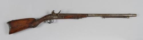 Fort fusil de chasse à silex, un coup. Canon rond, rayé, à pans au tonnerre, à point de mire décoré à l'or d'une toile d'araignée, gravé, poinçonné et enrichi à l'or au tonnerre de chutes de fleurs, de volutes, de foudres sur fond rayonnant et de la signature «ARMAND RUE DU ROULE A PARIS» (piqûres). Platine signée «Kivietniewski (?) Varsiawie» et chien à corps plat, gravés de drapeaux et feuillages, bassinet à deux volutes pare-étincelles. Lumière rehaussée d'or. Ressort de batterie à roulette. Entrée de baguette au masque d'Hermès en demi-ronde-bosse, gravée de poissons et passants bordés de frises parlées, en argent découpé. Contre-platine en argent découpé, ornée de dogues. Pontet à décor en demi-ronde-bosse de masque d'Hermès et de palmettes, en argent découpé et gravé d'urne fumante et de serpents. Plaque de couche en suite avec retour en demi-ronde-bosse (manque une vis). Crosse à joue et fût en noyer choisi, en partie quadrillé, sculpté de feuillages et d'une tête de dragon. Baguette en fanon à embout en laiton (cassée et en partie manquante). Poinçons de titre Coq 1, 950 et de moyenne garantie de Paris (1798-1809). A.B.E. (Accident mécanique). Époque Consulat - Premier Empire. Armand. Arquebusier à Paris (1780-1840). Vu sur un très beau fusil de chasse canons d'Henry Dombret. 268 puis 19 rue du Roule.