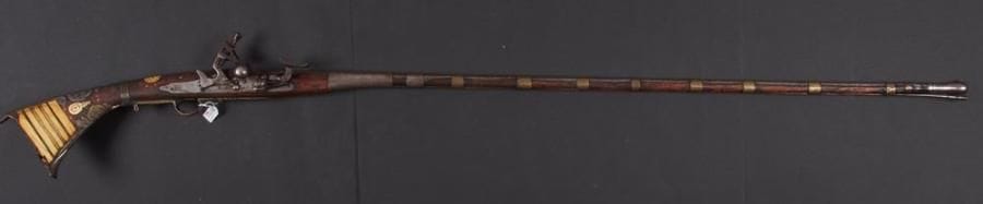 Fusil à silex d'Afrique du Nord dit Moukalah. Long canon rond, tulipé à la bouche, à pans au tonnerre avec bracelets en fer gravé et en laiton. Platine à la Miquelet. Crosse en noyer incrustée d'os, de laiton et de fer découpé à décor d'arabesques (petits manques). Baguette en fer. A.B.E. (Accident mécanique). XIXe siècle.
