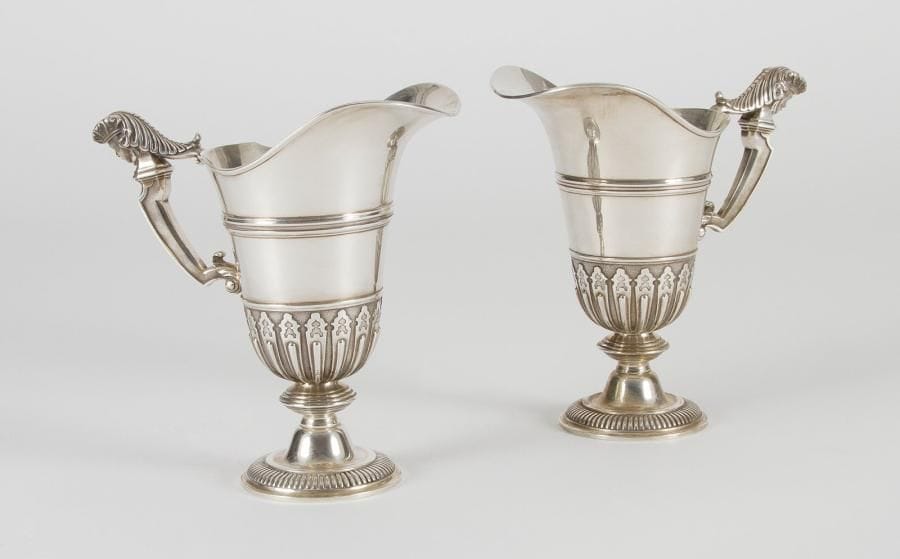Paire d'aiguières casque en argent, l'intérieur en vermeil, le piédouche à décor de godrons, le corps appliqué de lambrequins jusqu'à mi-corps sur fond amati, l'anse surmontée d'une tête de jeune homme coiffé, traité en ronde-bosse. Poinçon minerve. Orfèvre: TETARD frères. Poids: 2432 g Haut.: 24,2 cm