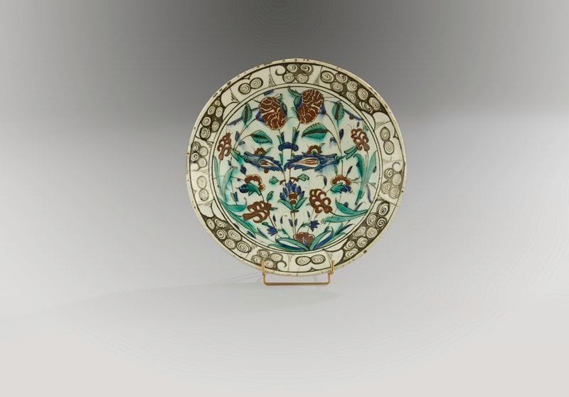 Plat tabak à bouquet symétrique, Turquie ottomane, Iznik, XVIIe siècle Céramique siliceuse à base annulaire à décor peint en polychromie sur fond blanc sous glaçure transparente. Composition symétrique d?un bouquet d?oeillets et de fleurs composites jaillissant d?une touffe d?herbe. Au rebord, frise de vagues et rochers noirs. Au revers, alternance de volutes en spirales bleues et vertes. Diam.: 30 cm et H.: 6 cm

An Iznik polychrome pottery dish, decorated with symmetrical flower motifs, Ottoman Turkey, 17th century(*)