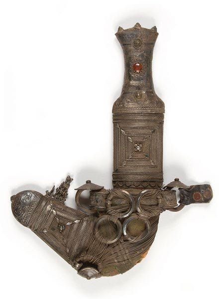 Poignard djambiya, Arabie, XXe siècle Lame courbe en acier à double tranchant et nervure médiane. Monture en argent à décor filigrané rehaussé de trois cabochons, dont un imitant la cornaline et coiffée de trois petits cônes. Fourreau en tissu à chape et bouterolle en argent à décor filigrané et perlé de carrés inscrits les uns dans les autres. Attache de boucles tenues par des fils d?argent, fixée sur le fourneau par un bandeau recouvert d?argent. Long.: 28 cm 


An Arabian horn-hilted dagger, Jambiyya, 20th century