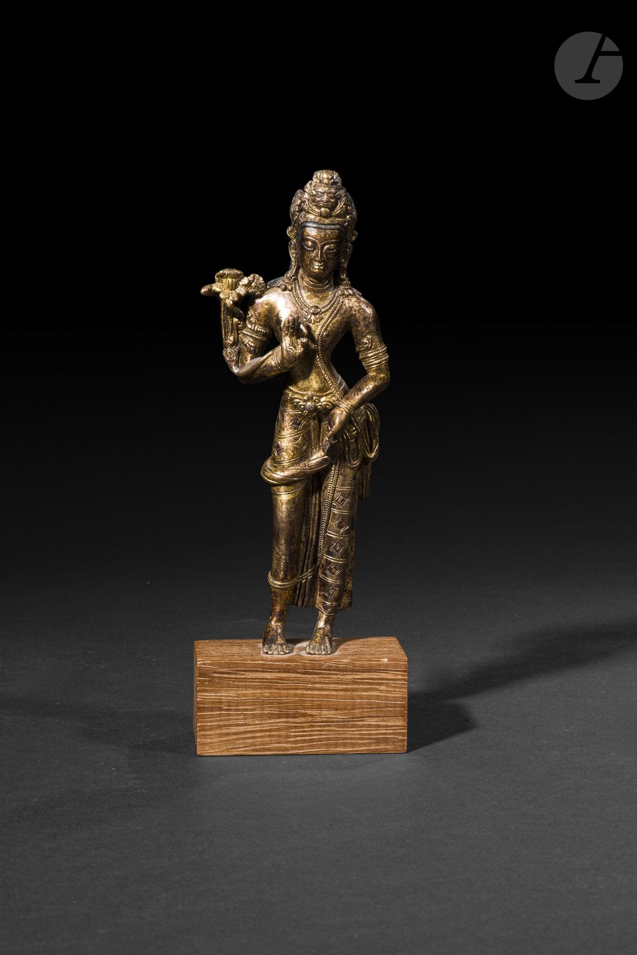 Statuette en alliage de cuivre doré représentant le bodhisattva Avalokitesvara (Padmapani), Népal ou Tibet, tradition himalayenne, XIXe - XXe siècle
Représenté debout en léger déhanchement (tribhaṅga), le bodhisattva adopte une attitude élégante et élancée. La main droite levée forme un vitarka mudrā, geste d’enseignement, tandis que la main gauche abaissée reprend une position analogue. Il est paré de riches bijoux princiers : colliers, bracelets et pendants d’oreilles et est vêtu d’un dhoti finement plissé retenu par une ceinture ornée, embellis d’incrustations d’argent. La tête est coiffée d’une couronne décorée d’un motif de kīrtimukha, visage serein aux yeux mi-clos. Un lotus épanoui, dont la tige repose le long de l’avant-bras droit et s’élève derrière l’épaule, constitue l’attribut iconographique caractéristique d’Avalokiteśvara sous sa forme Padmapāṇi, le bodhisattva de la compassion.
Hauteur : 17,3 cm