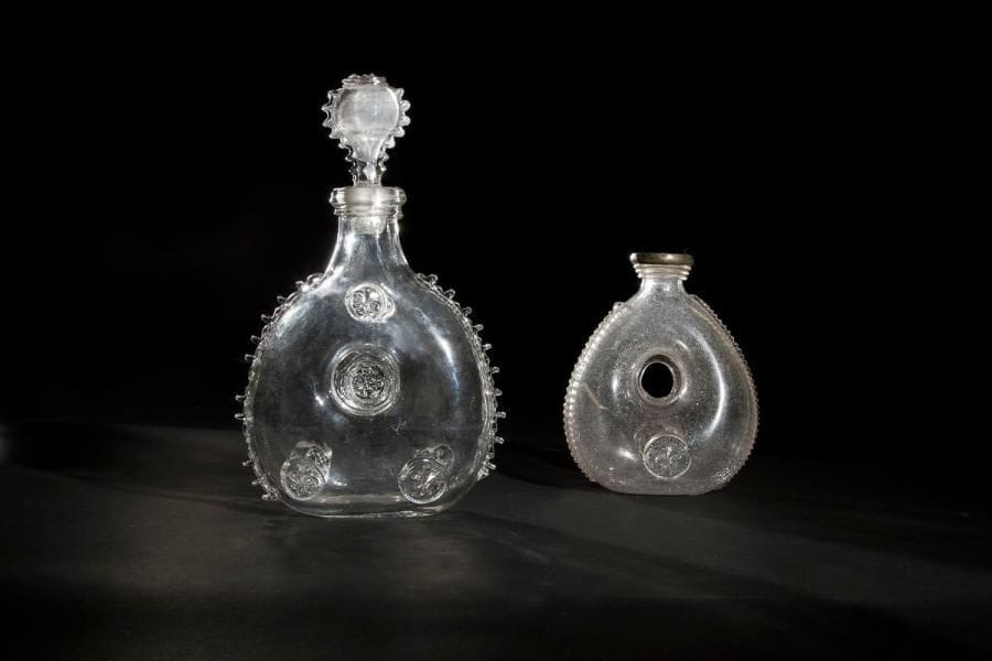 Deux gourdes annulaires en verre soufflé ornées en applique de crantages travaillés à la pince et de pastilles à motif de fleur de lys moulée: l'une, du XVIIIe siècle, le goulot cerclé d'étain (fêlure dans le corps), la seconde, dans le style du XVIIIe siècle, comporte un bouchon en verre soufflé (petite fêlure au col). Haut. 27,5 cm (avec bouchon) et 16 cm