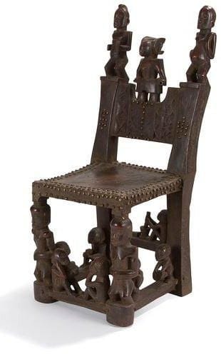 TSHOKWE (Angola) Chaise. Siège gainé de peau animale et orné de clous de tapissier en cuivre. Dossier sculpté de motifs géométriques et de 3 personnages dont le central présente une coiffe du type "mutwe wa kayanda" dérivant de la coiffure ailée du masque sacré "Cikungu" et indiquant le rang social très élevé. Les pieds et les traverses sont ornés de personnages et de scènes de la vie quotidiennes (proverbes?). Ce siège datant de la période coloniale 1940/1950 correspond aux sculptures de deuxième génération, les plus anciennes du XIXe siècle ayant été collectées ou détruites. Profondeur: - Hauteur: 70 cm - Longueur: 30 cm - Largeur: 35 cm Bibliographie: M. L. Bastin, La sculpture Tshokwé, Chaffin, 1982, p. 271 à 283