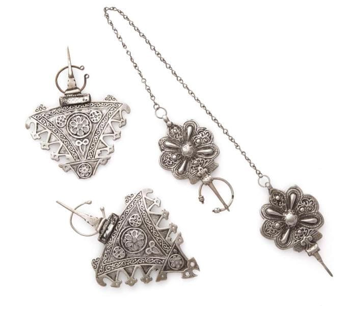 Deux paires de fibules en argent, Maroc, tribu du Haut Altas, Dadès, début XXe siècle Une paire de grandes fibules triangulaires à décor appliqué d'une grande rosette centrale flanquée de deux plus petites. Une autre paire alvéolée, moulée de rosettes cruciformes, reliées par une chaîne. Long.: 17,5 cm et 15,5 cm; 67 cm PB.: 162 g; 131 g État: manque le haut d'une fibule de la paire de grandes fibules; l'autre: manque un cercle. Two pairs of silver fibulae, Morocco, High Atlas, Dades Tribe, early 20th century