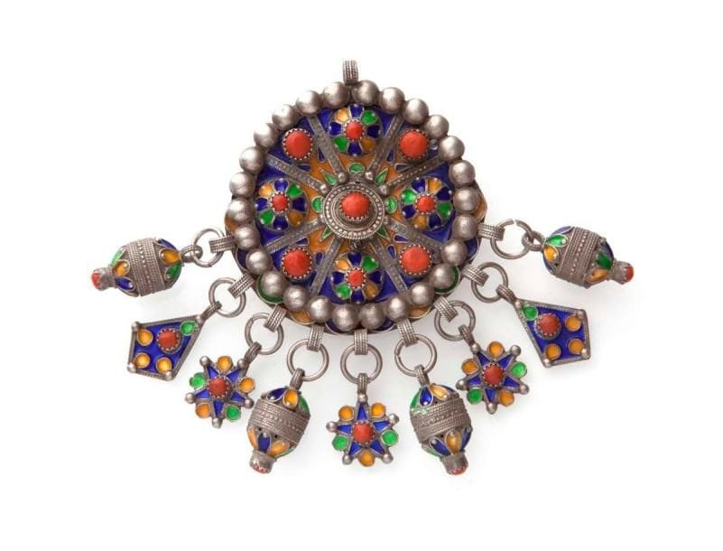 Fibule ronde en argent, Algérie, Tabzimt, Grande Kabylie, Beni Yenni, fin XIXe siècle-début du XXe siècle Argent ciselé, décoré d'émaux cloisonnés bleus, jaunes et verts, sur les deux faces, serti de huit cabochons de corail et d'un autre au centre sur l'ardillon de fermeture. Neuf pendeloques de trois types serties de corail et dos orné d'une rosace stylisée. H.: 114,5 cm (avec pendeloques) Diam.: 7 cm PB.: 130 g A round enamelled silver fibula, Algeria, Great Kabilya, Beni Yenni, late 19th- early 20th century