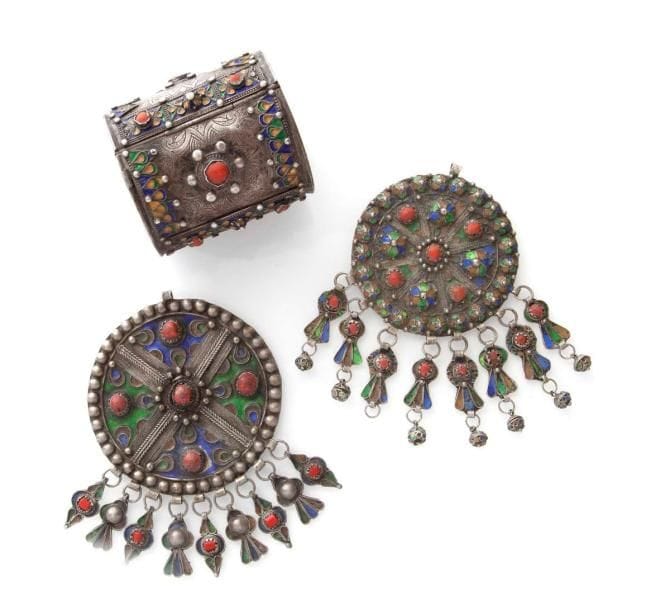 Chevillère (khelkhâl) et deux grandes fibules rondes en argent, Algérie, Grande Kabylie, Beni Yenni, début XXe siècle Argent ciselé, décor rehaussé d'émaux bleus, jaunes, verts. Chevillère avec charnière ouvrante. Fibules serties de quatre cabochons de corail et d'un autre au centre sur l'épingle de la fibule, agrémentées de huit et neuf pampilles serties de corail. H.: bracelet: 10 cm H. fibules avec pampilles: 17 cm; diam.: 12 cm et 10 cm PB.: bracelet: 316 g; fibules: 196 g et 160 g État: manque une pampille sur une fibule. Pour des modèles similaires, voir: Camps Faber H., Bijoux berbères d'Algérie, Edisud, 1990. A silver anklet (Khelkhâl) and two large round enamelled silver fibulae, Algeria, Great Kabylia, Bani Yenni, early 20th century