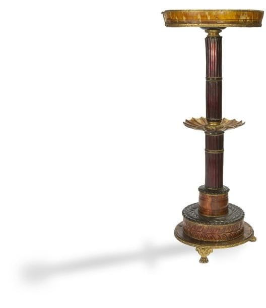 Rare petit guéridon en écaille brune et rouge, corne, loupe d'amboine et bronze doré, le plateau en cabaret reposant sur un fût cannelé et bagué incorporant une coupe à décor de velum et reposant sur une base circulaire à emmarchements à décor en relief et pieds griffes à volutes.
Travail probablement anglais d'époque Regency (Restaurations).
H. : 72 cm - L. : 28,5 cm
D'allure assez légère, ce guéridon reprend de nombreuses caractéristiques du mobilier de l'époque Regency notamment le fût segmenté de bagues reposant souvent sur le même type d'entablement à pieds en griffe.
Le décor présente également une stylisation du motif feuillagé traité en relief, notamment au piétement, sur le dessus ainsi qu'à la circonférence de la base, qui évoque aussi certains guéridons anglo-indiens. De plus, le motif de velum apparaît comme un leitmotiv dans le mobilier anglais en général.
L'usage de l'écaille est par ailleurs parfaitement avéré en Angleterre et aux Indes britanniques dans le courant du XIXe siècle. On répertorie notamment de nombreux coffrets en écaille reposant sur des pieds en griffe