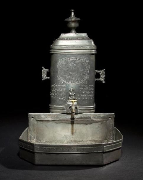 [GISSEF] FONTAINE ET SON BASSIN Allemagne, XVIIIe siècle, datée en hébreu 1783. Étain fondu et gravé. Fontaine: 30 x 19,5 cm Bassin: 9 x 25,8 x 25,5 cm 
Fontaine complète de tous ses accessoires: son couvercle, son robinet et son bassin. Elle porte gravé dans un encadrement circulaire sur sa face avant, une scène montrant Moïse tapant de son bâton sur le rocher pour en obtenir de l'eau. Elle est gravée en hébreu sur le couvercle de la date 543 du petit comput [1783]. Sur sa face avant et à droite du robinet, elle est gravée en hébreu du verset suivant:[Moïse leva la main, il frappa le rocher de son bâton par deux fois; il en sortit de l'eau en abondance (Nombres 20, 11).] Sur sa face avant et à gauche du robinet, elle est gravée du texte suivant:[J'épancherai sur vous des eaux pures afin que vous deveniez purs; de toutes vos souillures et de toutes vos abominations, je vous purifierai.] Mots-clés: Judaica, Jewish, Hebrew, Judaïsme, Hébreu.
(GISSEF) Ewer and Bowl. Germany, 18th century, dated in Hebrew 1783. Cast engraved pewter.