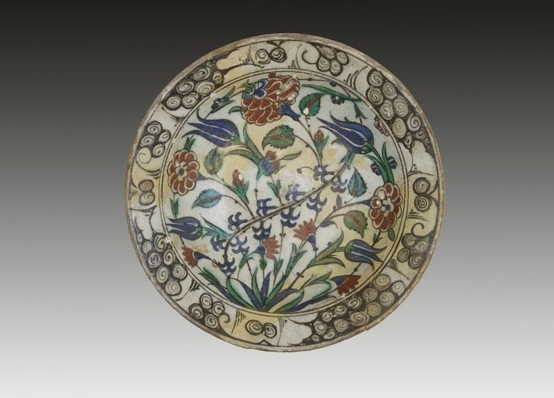 Plat tabak au bouquet, Turquie ottomane, Iznik, XVIIe siècle.
Plat creux sur base annulaire, en céramique siliceuse, peint en polychromie bleu, vert, rouge et noir, sous glaçure transparente, à décor central de tiges ondulantes d'?illets, tulipes et jacinthes. Au rebord motif de «vagues et rochers» soulignés de noir.
Diam.: 29 cm 
État: morceaux recollés, nombreuses restaurations
An Iznik polychrome pottery dish, decorated with flower motifs, Ottoman Turkey, 17th century
