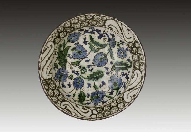 Plat tabak aux fleurs bleues, Turquie ottomane, Iznik, XVIIe siècle.
Plat creux à base annulaire, en céramique siliceuse, peint en polychromie sur fond blanc et sous glaçure transparente. Composition en plein d'enroulement de tiges de palmes saz et de fleurs hatay. Au rebord, frise de «vagues et rochers» stylisés en s et fleurs stylisées au revers. Trous de suspensions à la base. 
Diam.: 29,4 cm 
État: éclats de glaçure 
An Iznik polychrome pottery dish, decorated with blue flowers, Ottoman Turkey, 17th century