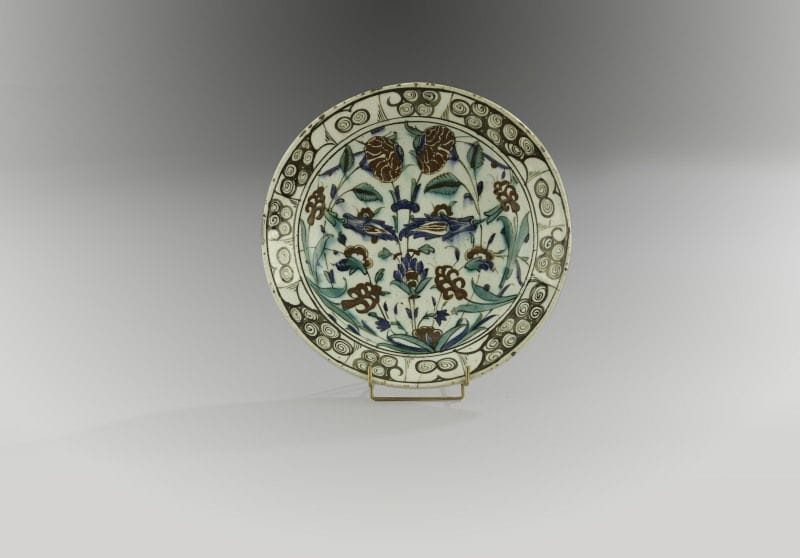 Plat tabak à bouquet symétrique, Turquie ottomane, Iznik, XVIIe siècle.
Céramique siliceuse à base annulaire à décor peint en polychromie sur fond blanc sous glaçure transparente. Composition symétrique d'un bouquet d'?illets et de fleurs composites jaillissant d'une touffe d'herbe. Au rebord, frise de «vagues et rochers noirs». Au revers, alternance de volutes en spirales bleues et vertes 
Diam.: 30 cm, Haut.: 6 cm
An Iznik polychrome pottery dish, decorated with symmetrical flower motifs, Ottoman Turkey, 17th century(*)