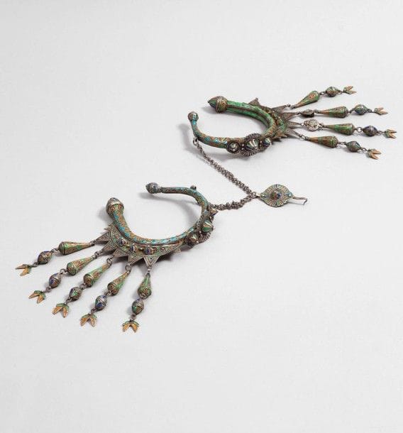 Paires de boucles temporales, Maroc, Anti-Atlas occidental, XXe siècle.
Deux larges anneaux reliés par une double chaînette et attache à crochet, à décor filigrané émaillé bleu, jaune, vert et turquoise d'un croissant dentelé et cabochons pyramidaux appliqués, à collerettes de rosaces pour certains, pendeloques coniques à pampilles boules.
Diam.: 15 cm 
P.B: 422 g
A paire of head ornaments, Morocco, Anti Atlas, 20th century