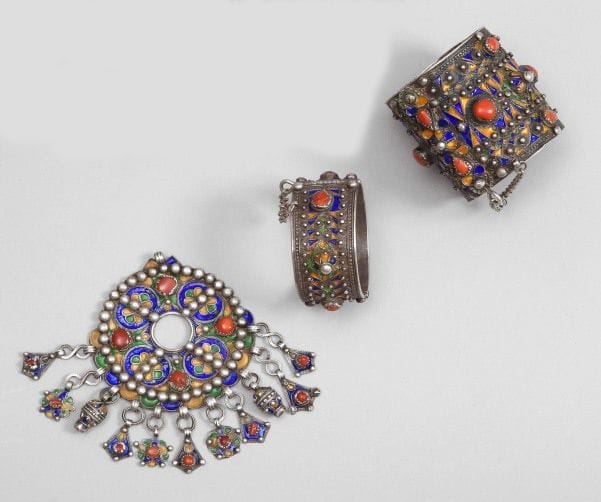 Fibule tabzimt, et deux bracelets, Algèrie, Grande Kabylie, Beni Yenni, XXe siècle. 
Fibule ronde en argent, émaillée sur fond vert et jaune de cercles bleus à c?ur floral jaune et vert alternant avec des cabochons de corail, bordée de perles appliquées ; - Bracelet manchette et bracelet articulés à décor émaillé vert jaune et bleu, cabochons en dôme et de corail, perles appliquées. 
Diam. fibule: 5.5 cm ; bracelets: 8 cm
P.B. fibule: 206 g ; bracelets: 106 g
An enamelled silver fibulae and two enamelled silver bracelets, Algéria, Great Kabylia, Bani Yenni, early 20th century